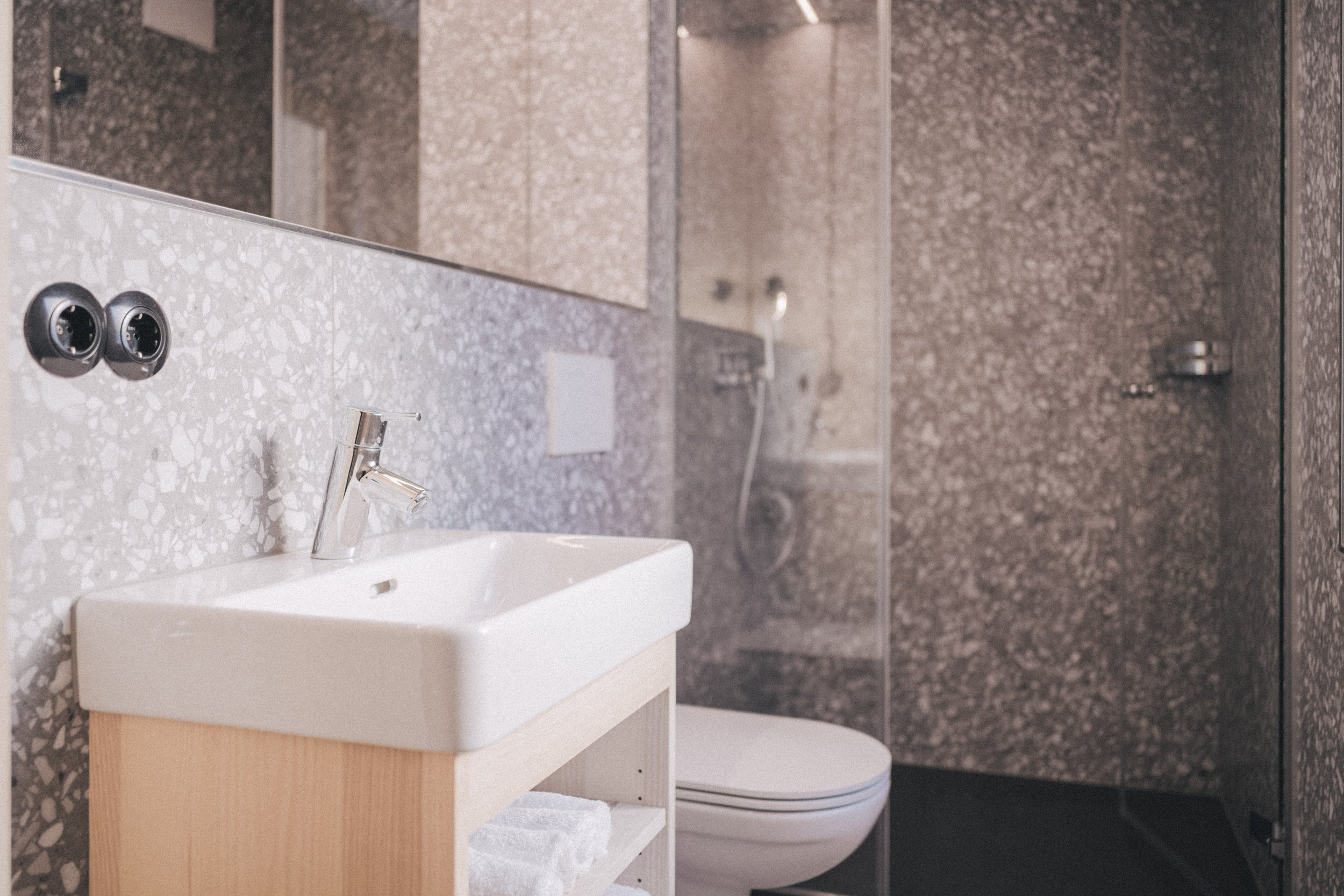 Ein Badezimmer mit eine kleine Waschbecken, eine Armatur, einem Spiegel, einem Handtuch und einer Toilette im Hintergrund. Wände und Boden sind mit schwarzem und grauem Mosaikfliesen bedeckt.