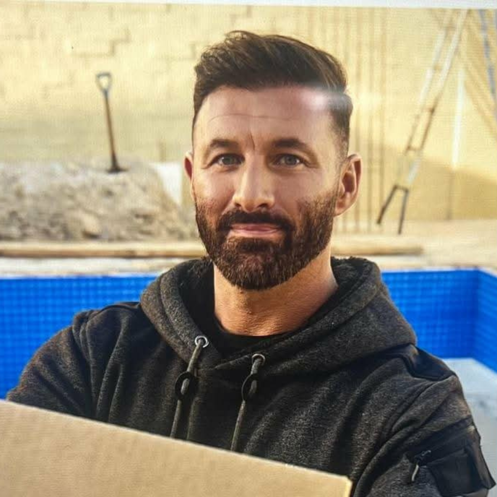A man with a beard and short hair taking a selfie on a construction site or backyard with a shovel and building materials in the background.