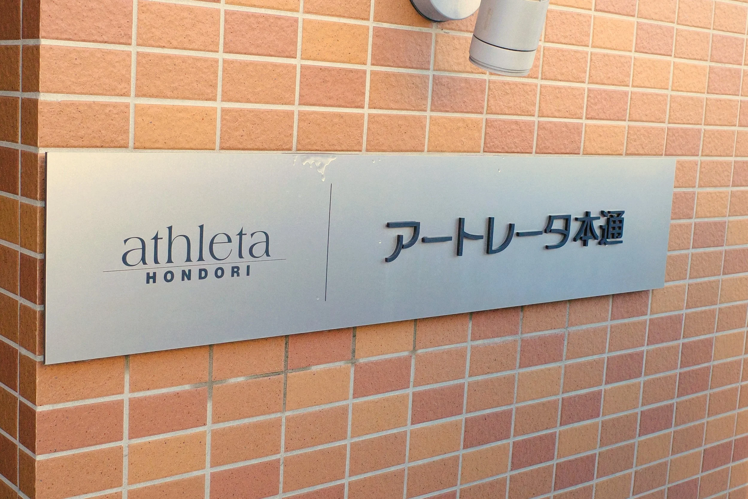 レンガの壁に設置された看板と照明。看板には「athleta HONORI」と書かれ、その横に日本語で「アートレータ本通」と記されている。