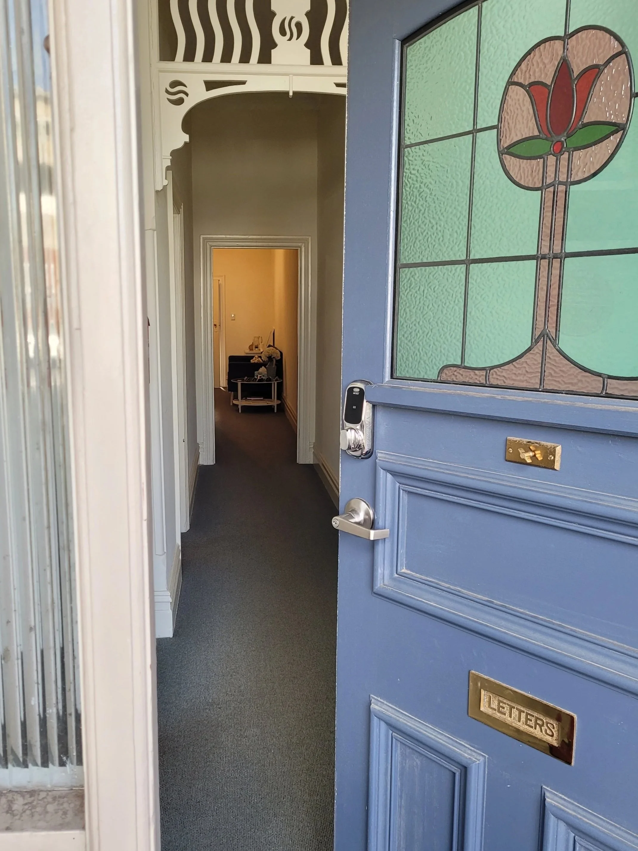 View of the entrance door of Lifeflow Psychology with a stained glass window featuring a red flower design, a brass mail slot labeled 'LETTERS,' a digital door lock, and a glimpse into a hallway with a small table and decorative items.