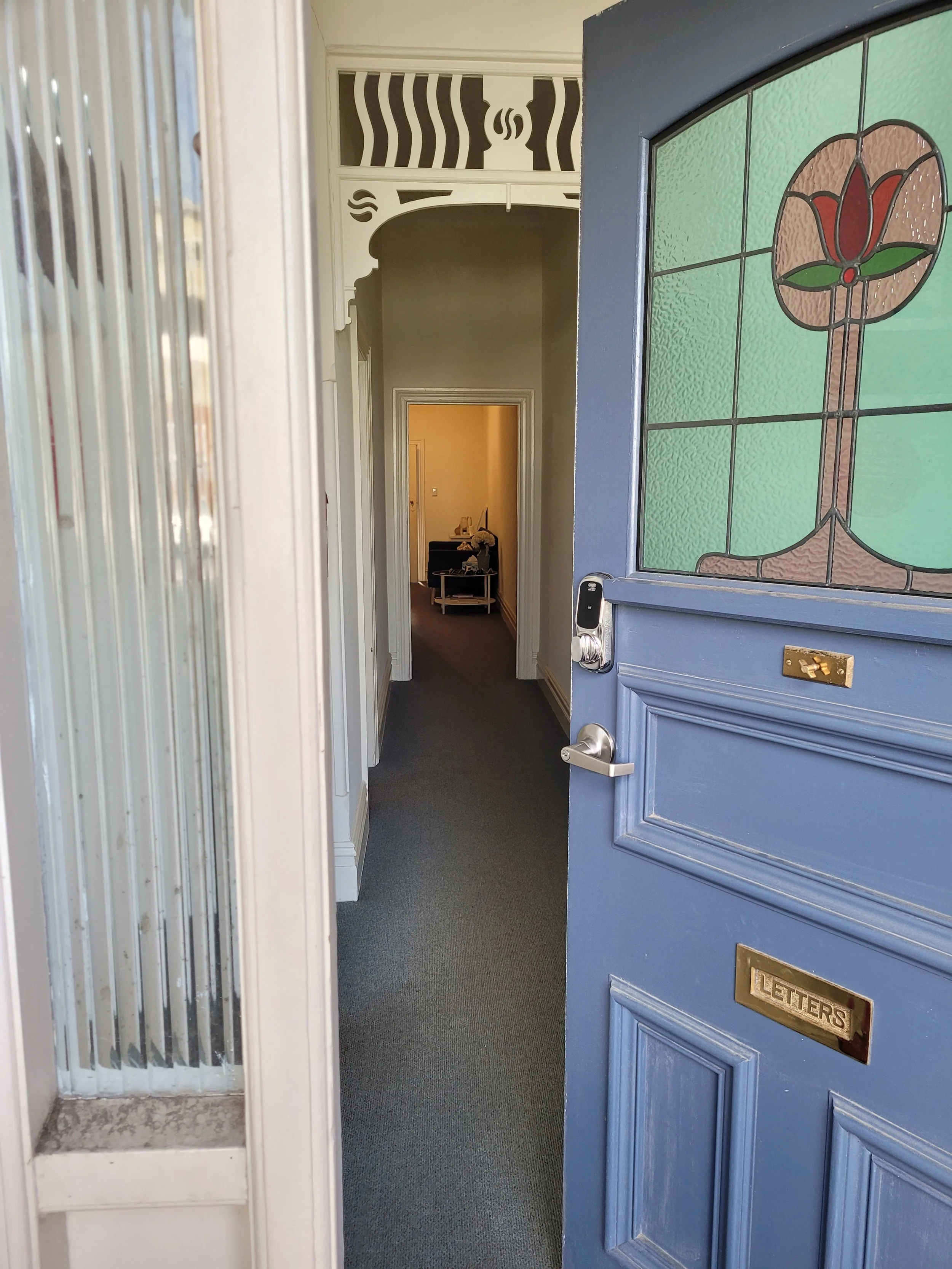 View of the entrance door of Lifeflow Psychology with a stained glass window featuring a red flower design, a brass mail slot labeled 'LETTERS,' a digital door lock, and a glimpse into a hallway with a small table and decorative items.