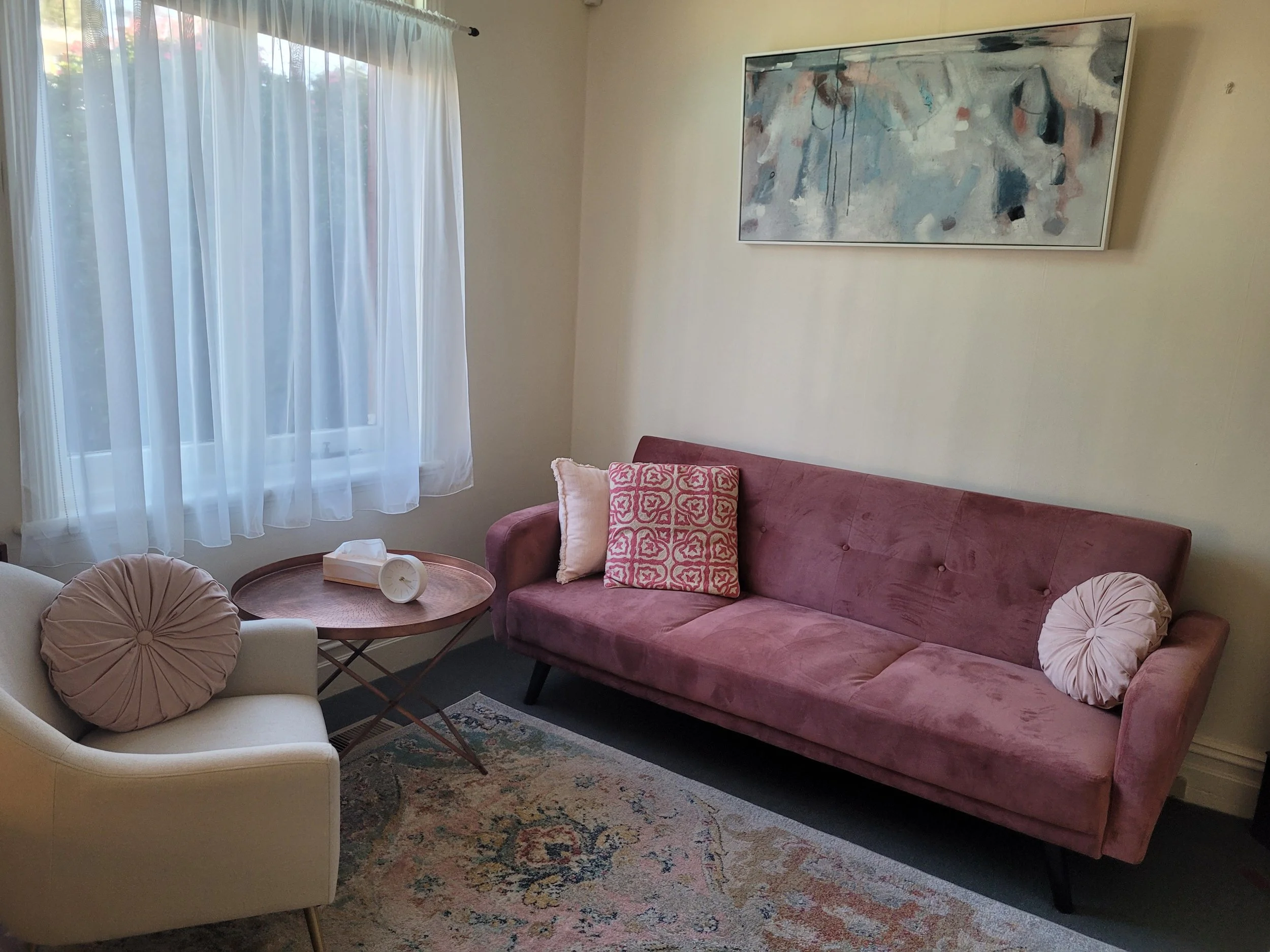 Clinic room at Lifeflow Psychology with a pink velvet sofa, beige armchair with round pillow, a wooden coffee table with tissue box and clock, abstract painting on the wall, large window with sheer white curtains..