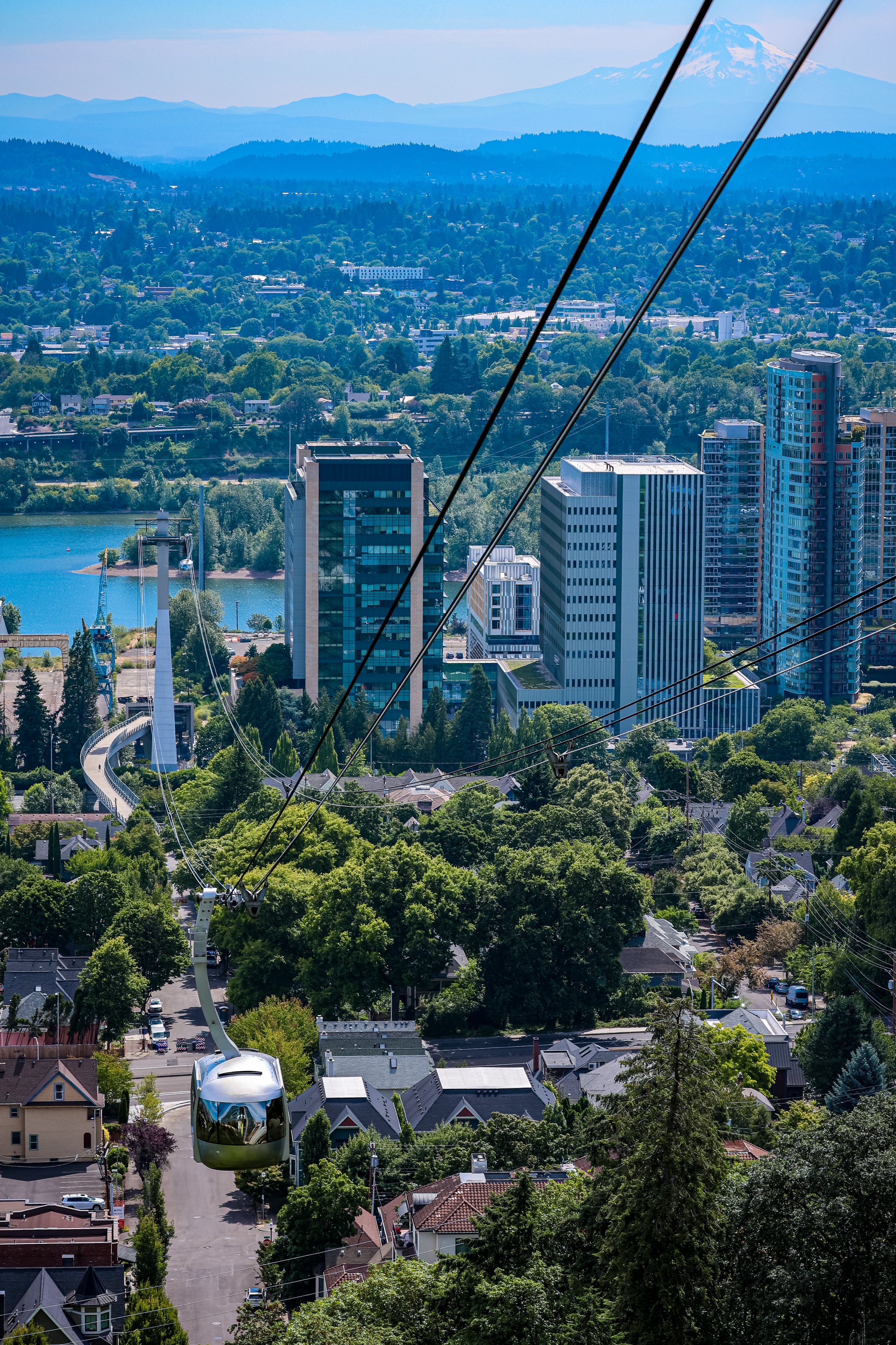 Aerial view of a cityscape with modern high-rise buildings, a river, and greenery. In the background, Mount Rainier is visible with snow on its peak. A large Ferris wheel and cable car are in the foreground.