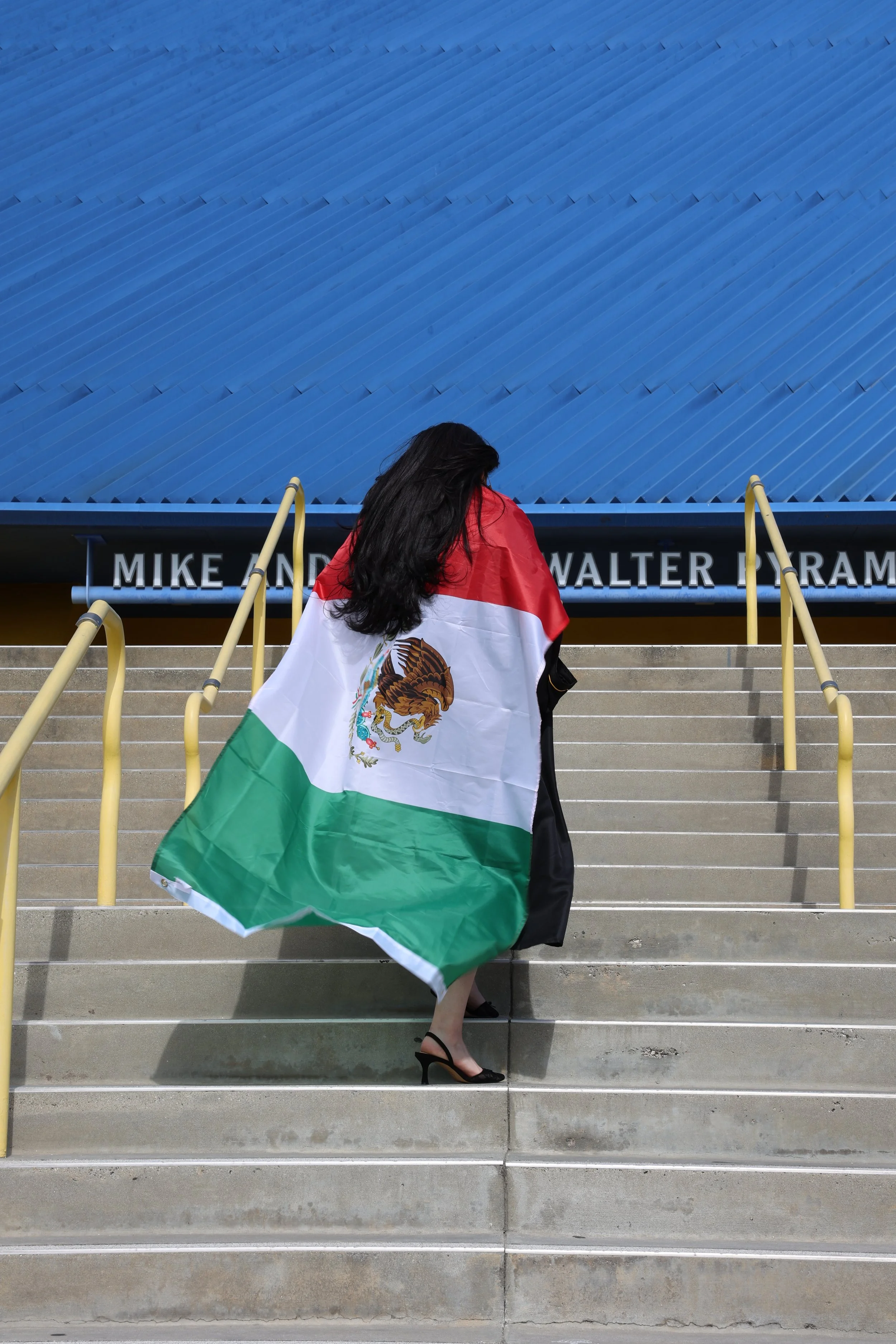 A woman standing on outdoor stairs with a Mexican flag draped over her shoulders. The stairs have yellow handrails and a sign at the top reads 'Mike and Walter Pyram'. She is wearing black high heels and a black dress, with her long dark hair covering her face.