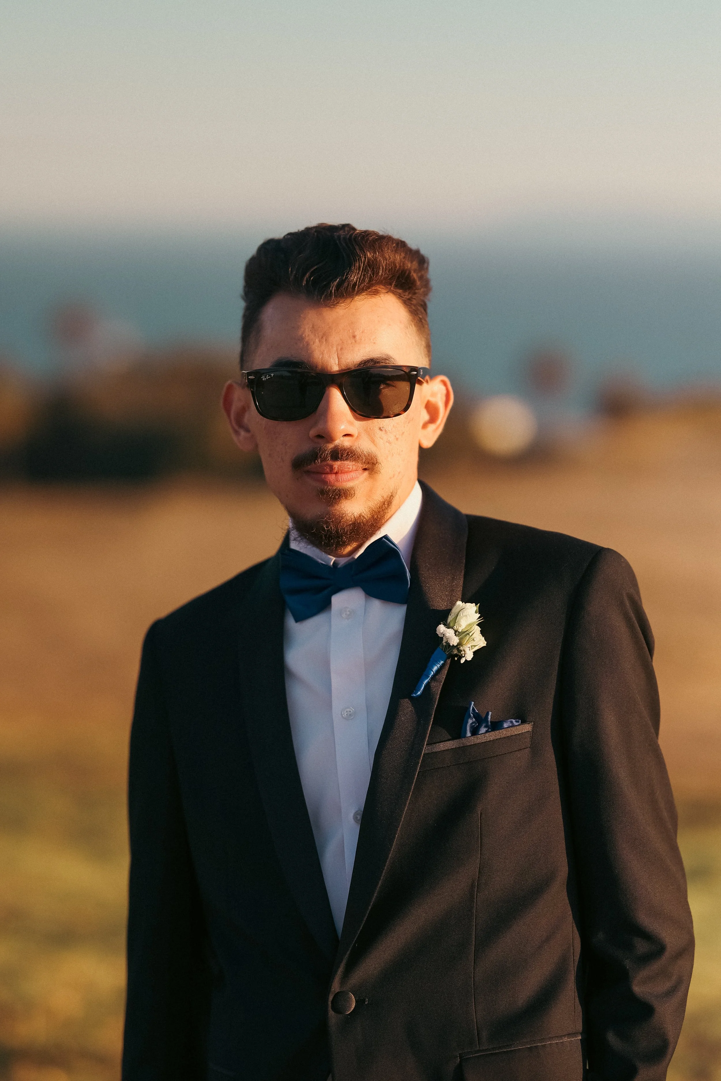 A man in a tuxedo with a boutonniere, dark sunglasses, and styled hair standing outdoors at sunset or sunrise.