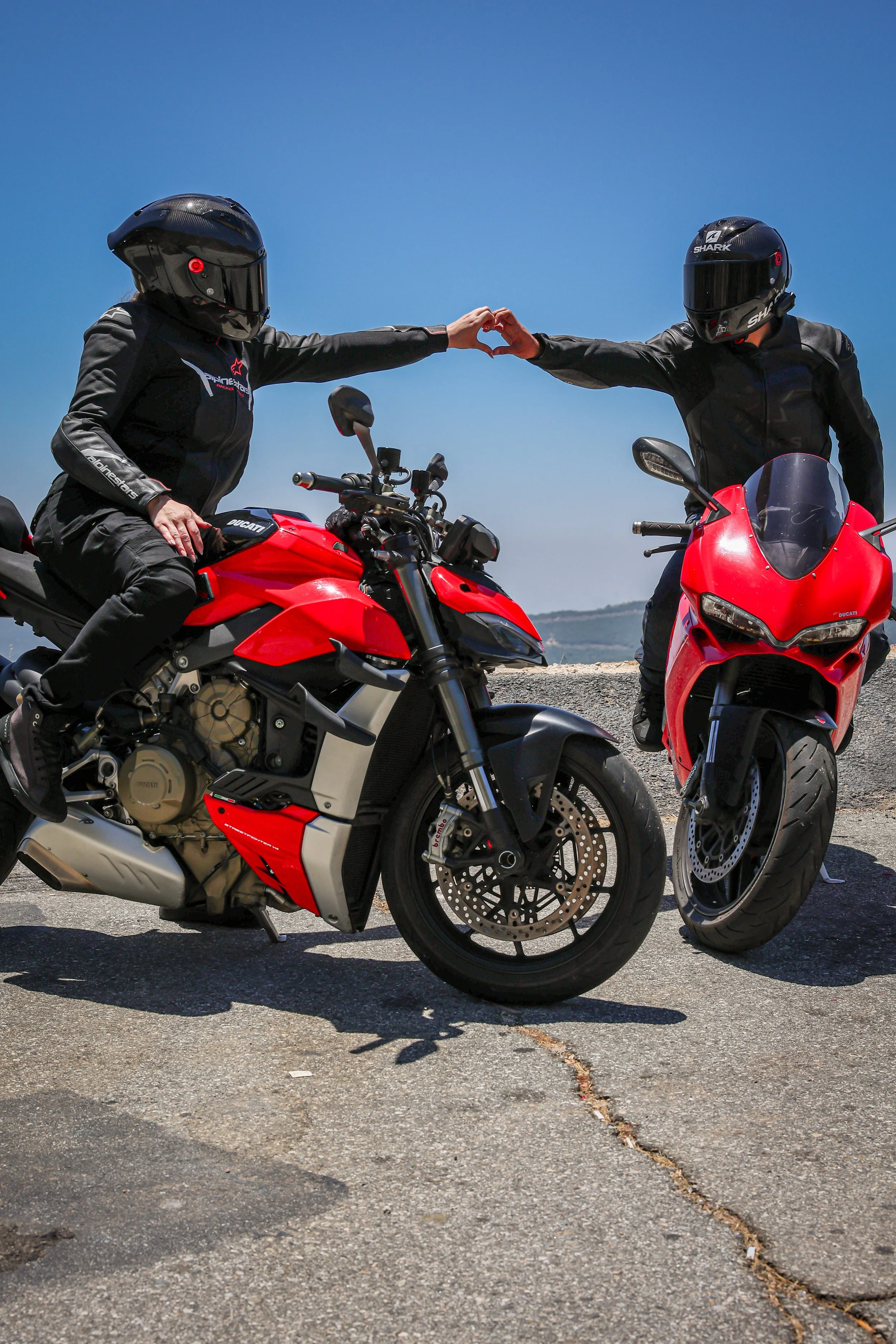 Two motorcyclists wearing black helmets and black riding gear sitting on red motorcycles, forming a heart shape with their hands against a clear blue sky.
