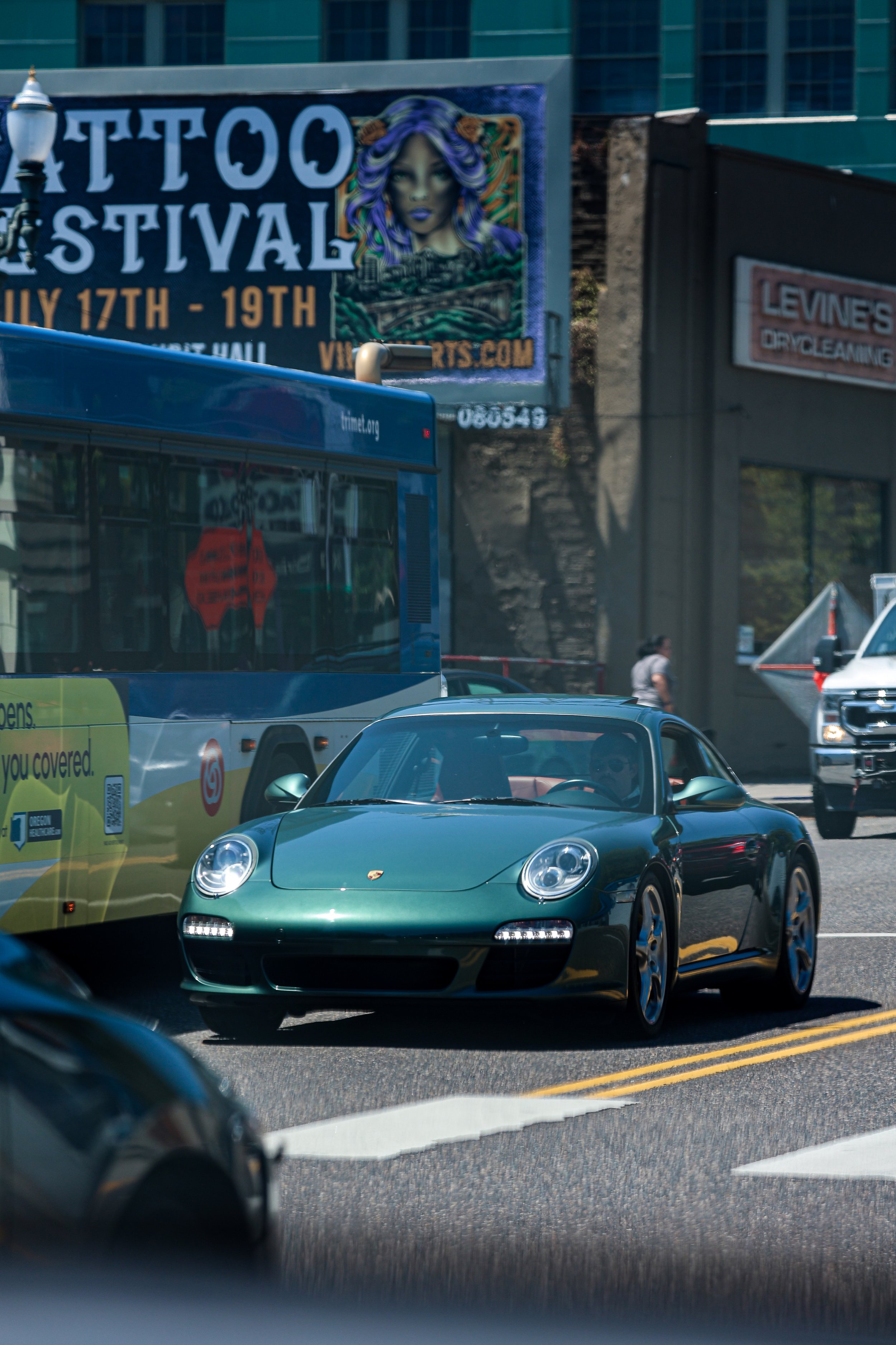 A black Porsche sports car on a busy city street with buses and pedestrians, with a billboard in the background advertising a tattoo festival from July 17th to 19th.