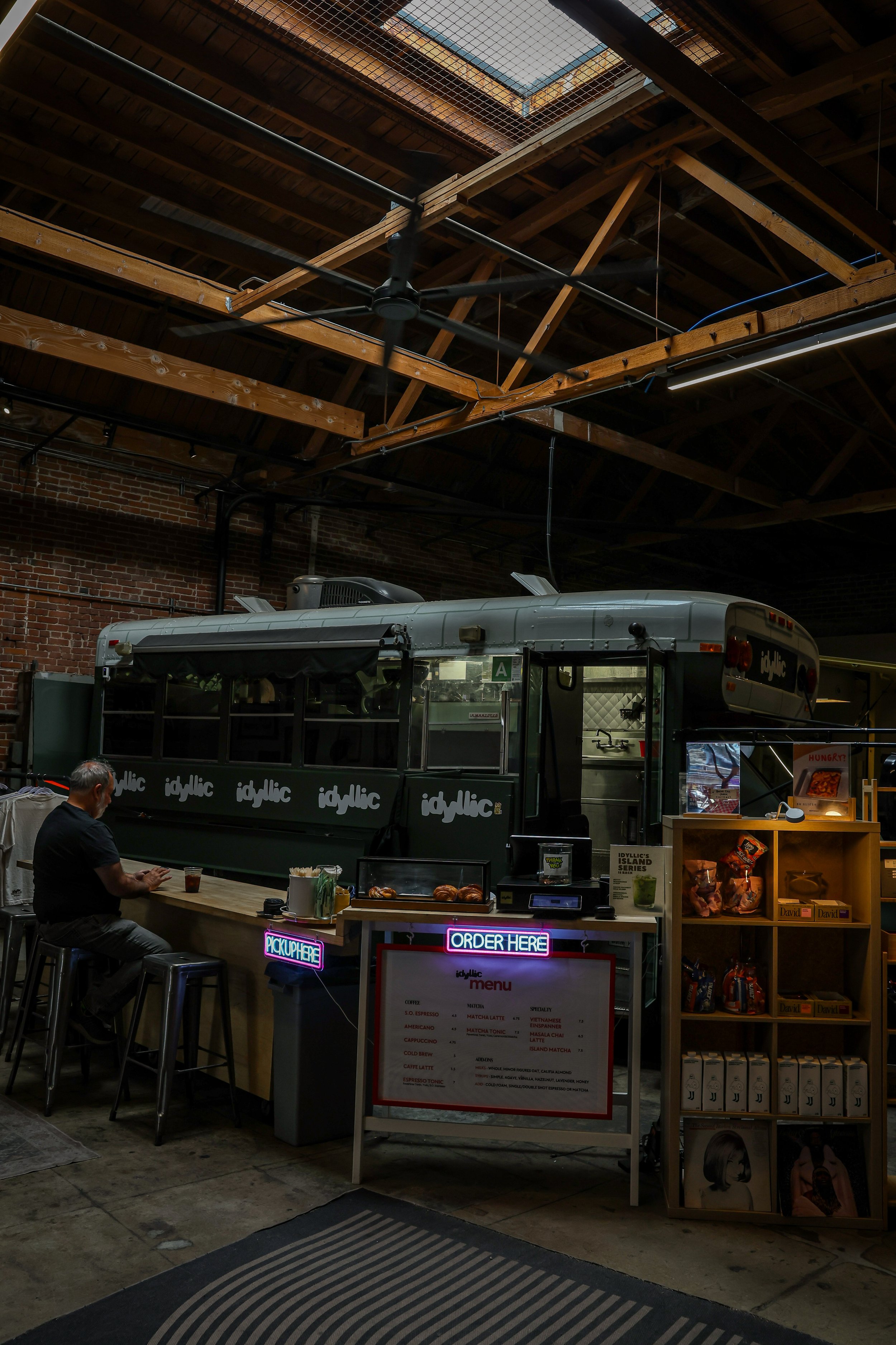 Inside a cafe or food stall with a vintage bus as the counter. A man is sitting at a bar-style table, drinking. The menu is displayed on a whiteboard, and a small lit sign reads 'Order Here.' Shelves with snacks and decorative artwork are visible on the right. The ceiling has exposed wooden beams and a ceiling fan.