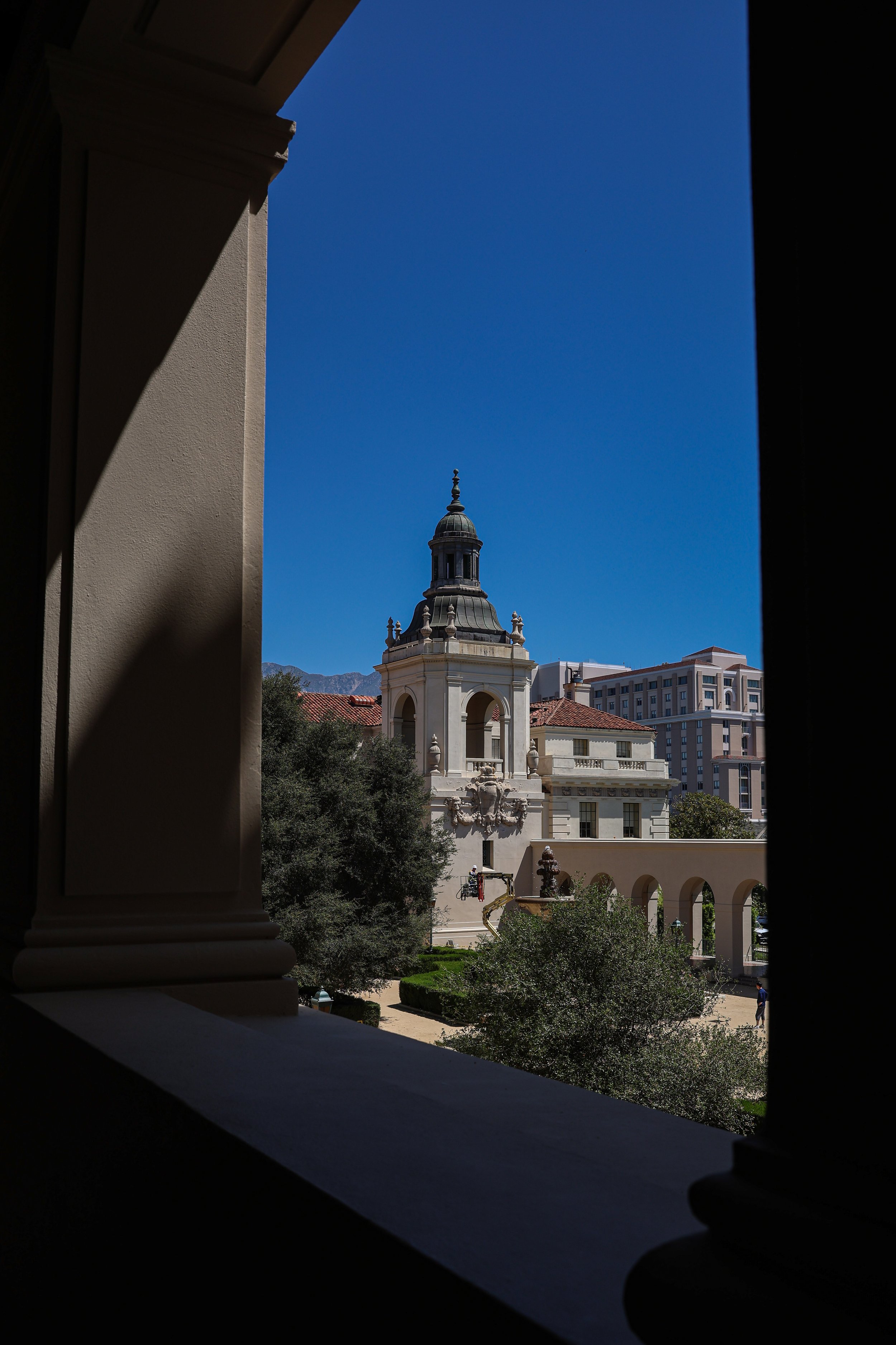 View of a historic building with a domed tower and a fountain in front, seen through a window with columns and a ledge, with mountains and modern buildings in the background under a clear blue sky.
