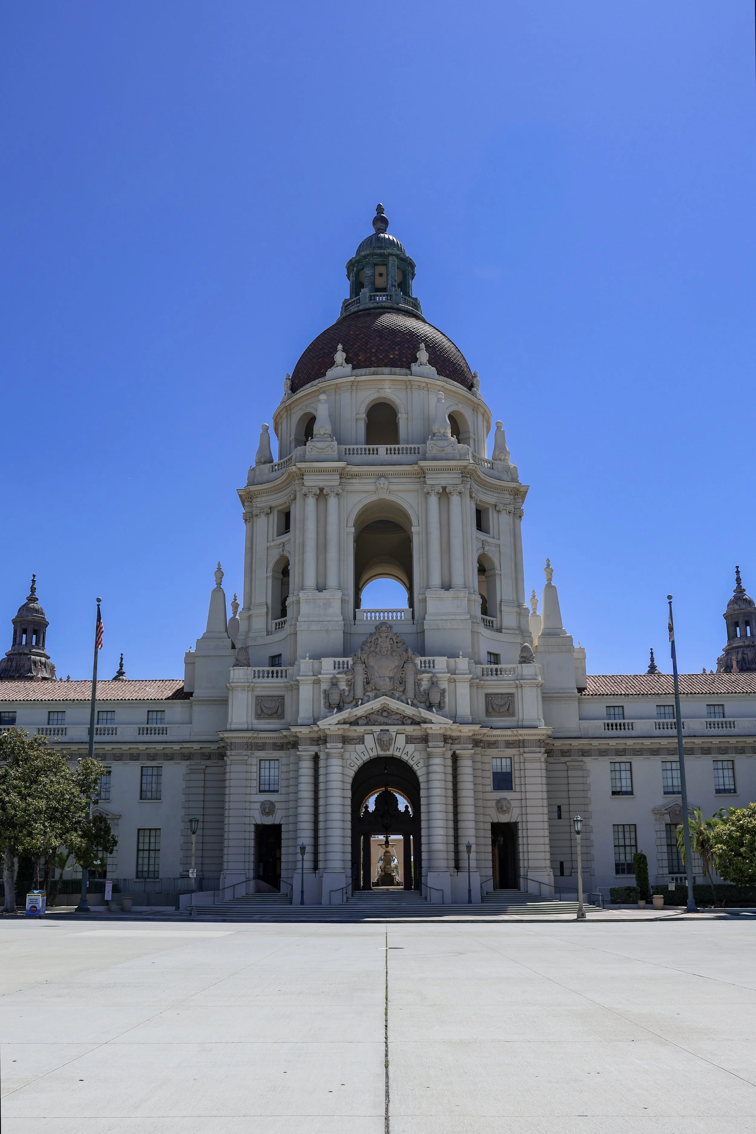 The image shows a historic city hall building with a large dome on top, ornate architectural details, and multiple archways and windows. The building is white with a red tile roof and is set against a clear blue sky. There are a few flags and some trees in the foreground.