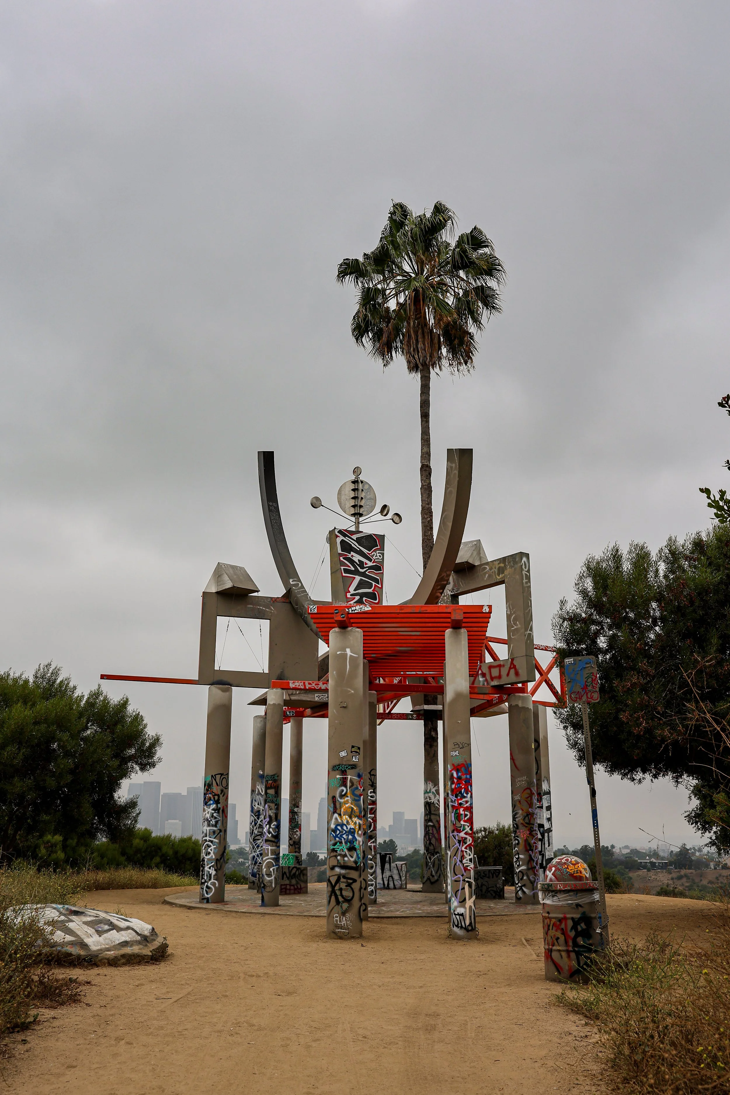 A tall palm tree with a thin trunk and green fronds stands behind a modern sculpture structure made of cylindrical concrete pillars covered in graffiti. The sculpture features curved metallic elements and a sign with graffiti. The area has dirt paths, some shrubbery, and a distant city skyline under a cloudy sky.