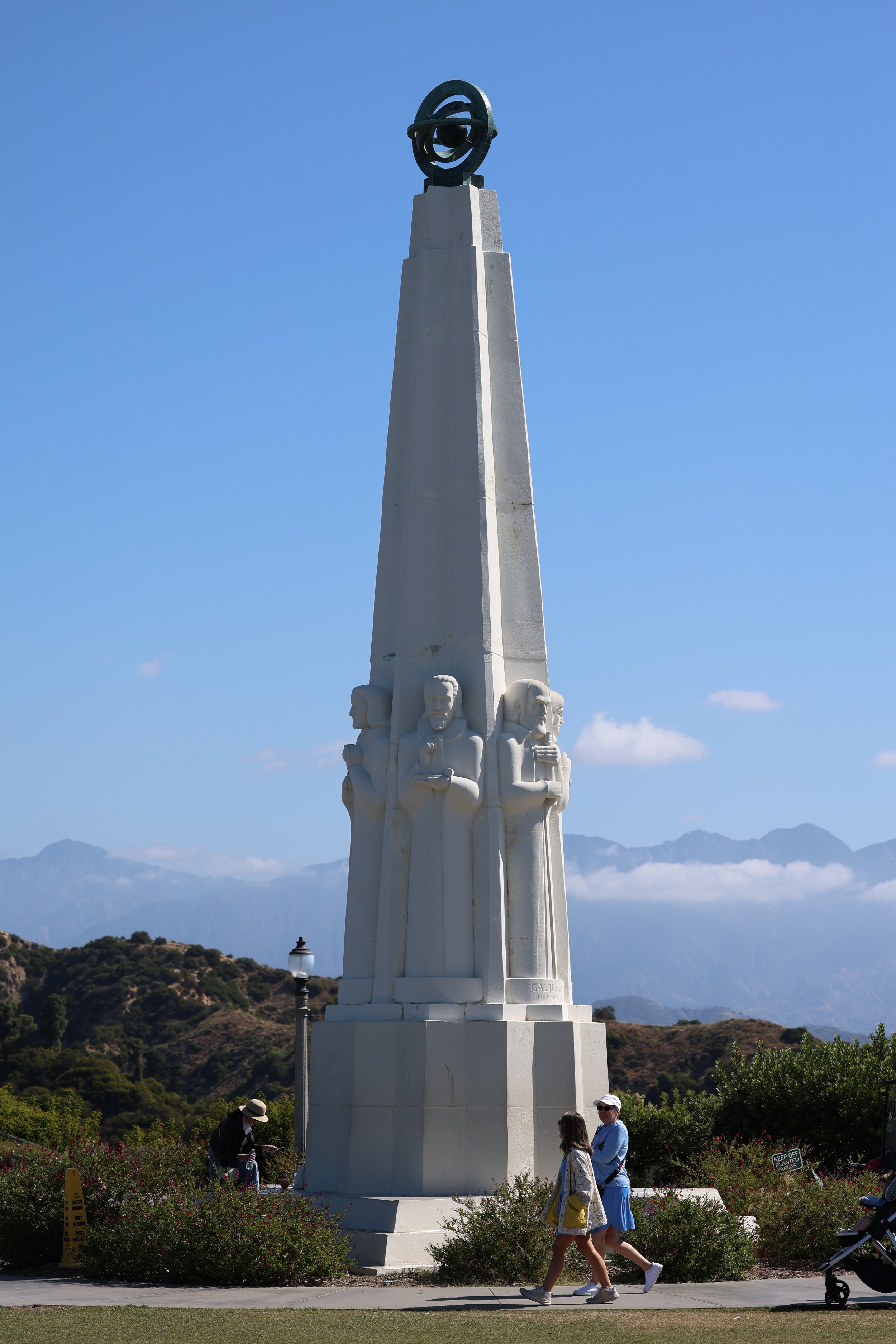 A large white statue featuring multiple carved faces at the base and a tall pillar with an abstract ring sculpture at the top, set against a clear blue sky with mountains in the background. Several people are walking and observing the monument outdoors.