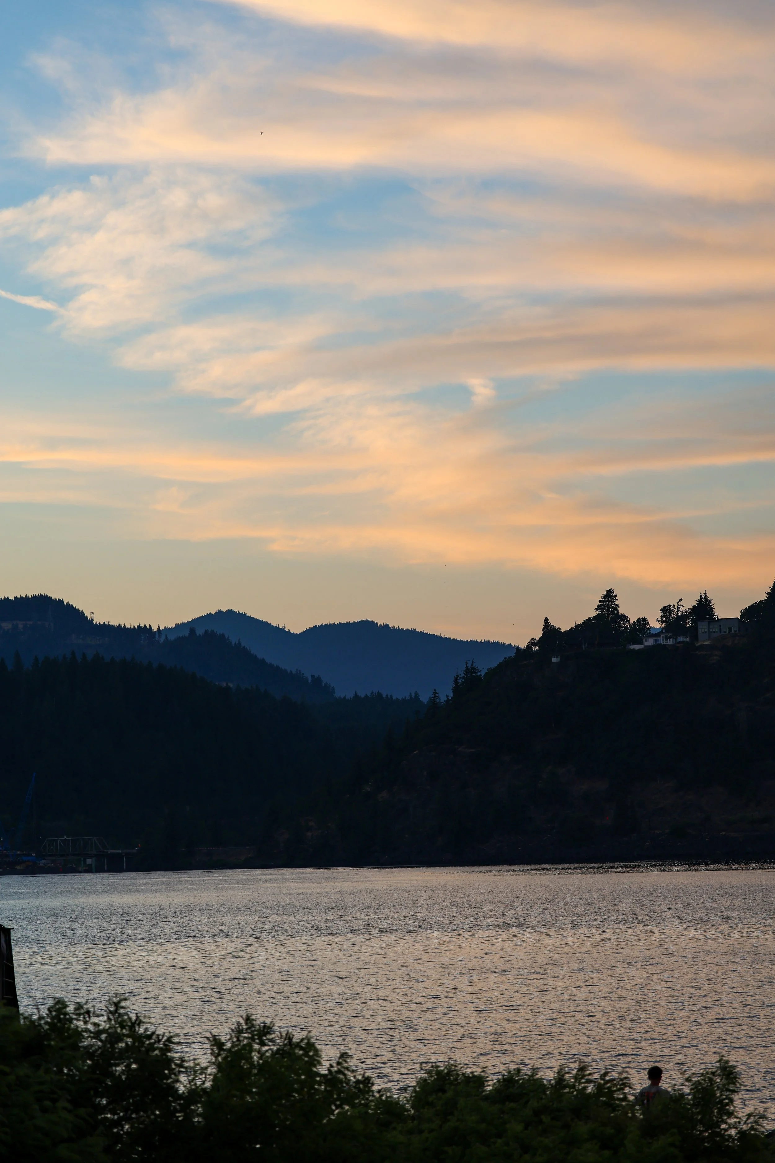 Scenic view of a body of water at sunset with mountains in the background and trees in the foreground, and a person standing near the water's edge.