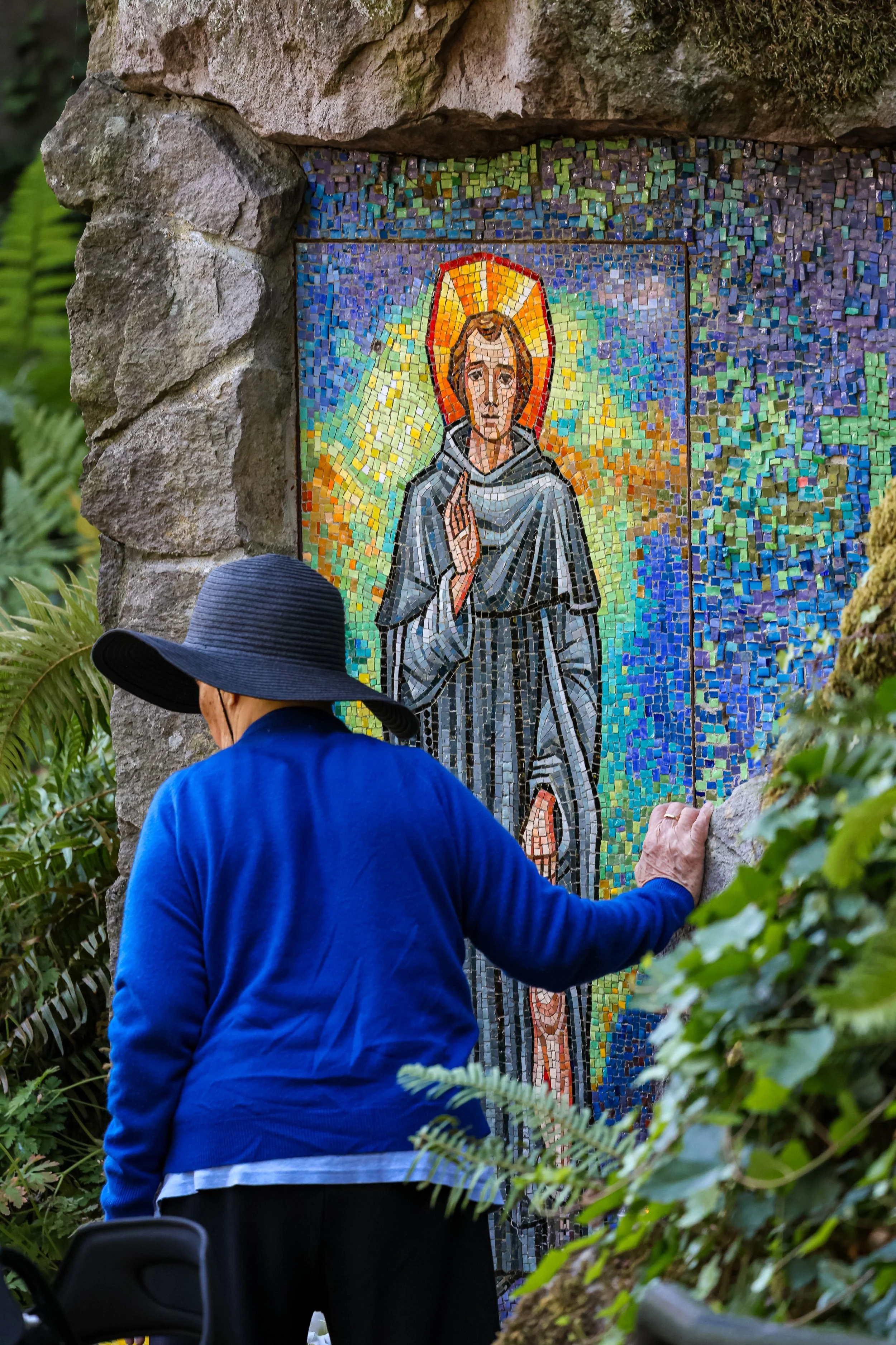 A person with a blue jacket and large black hat touching a mosaic of Jesus Christ, which is surrounded by greenery and set in a stone frame.