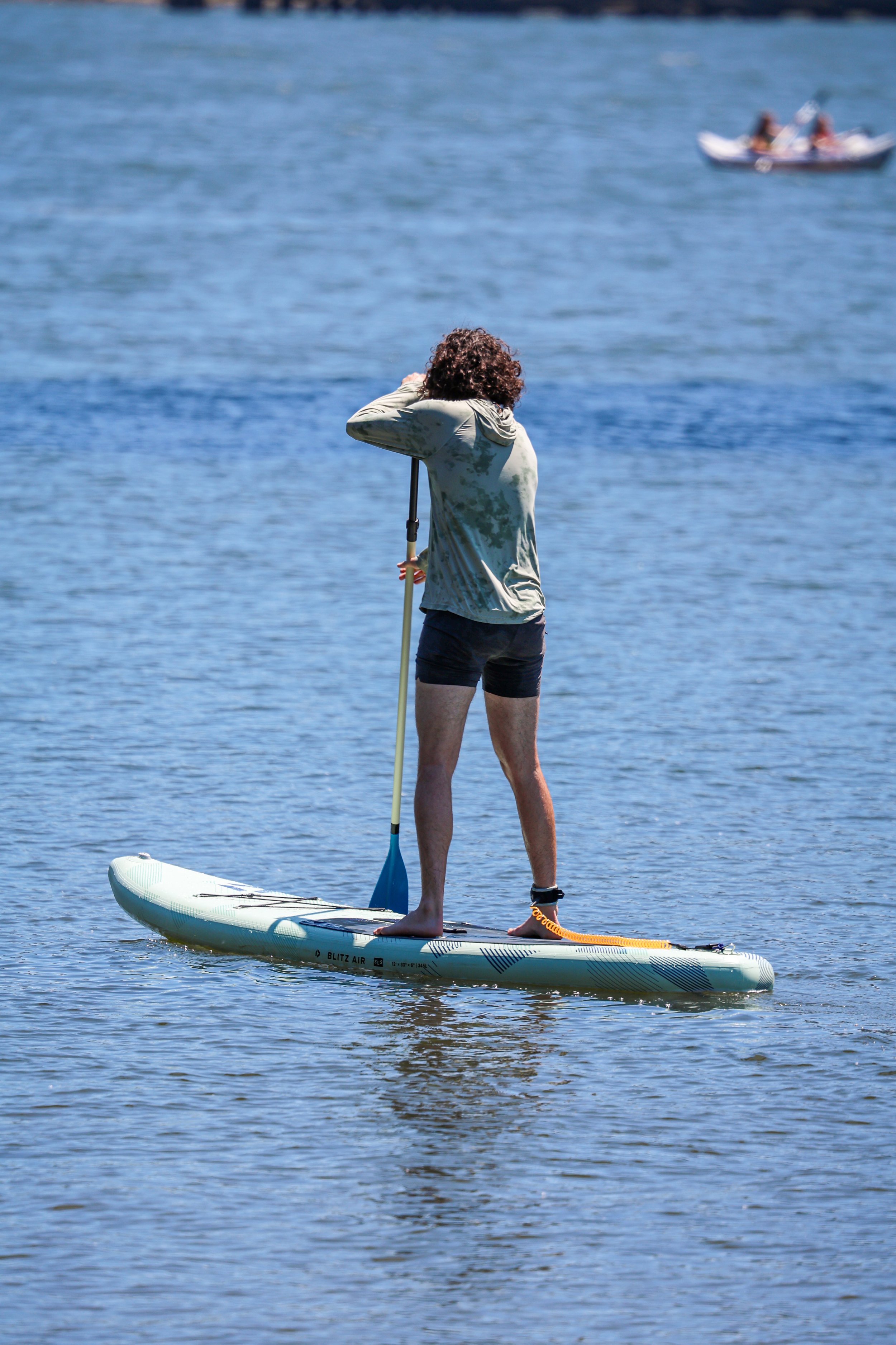 Person with curly hair stand on paddleboard on calm water, holding paddle and looking away from camera, with a boat and people in the background.
