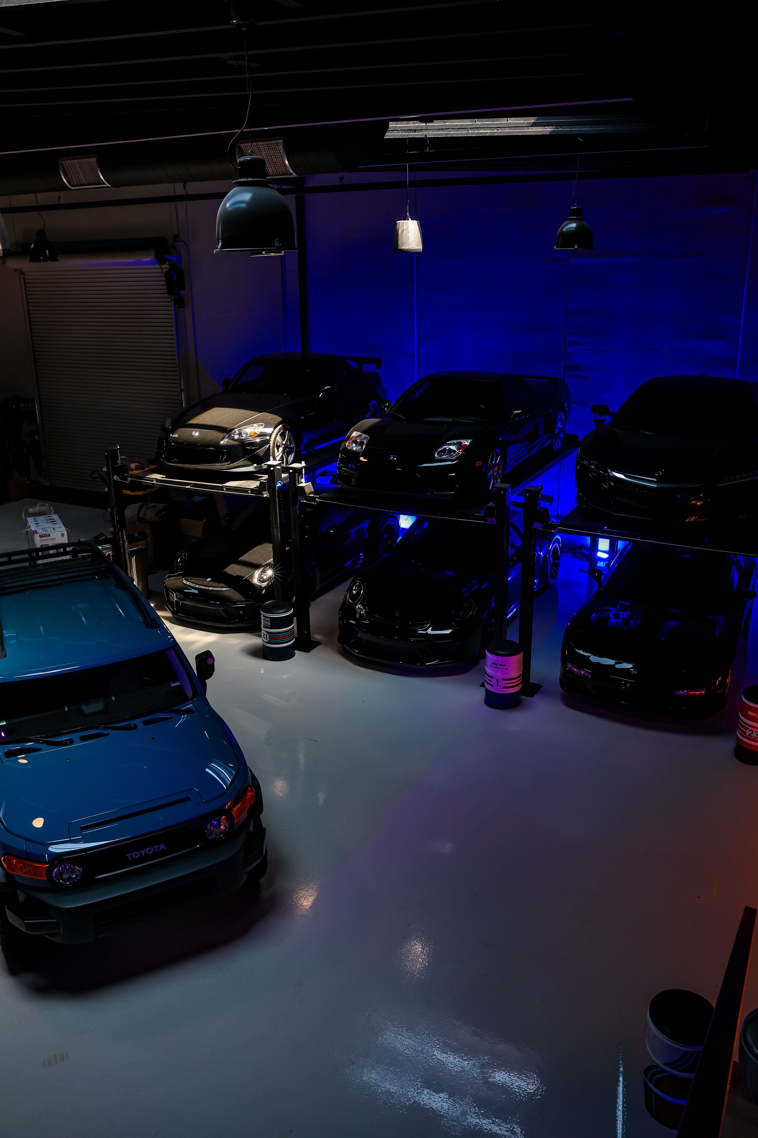 A collection of black and blue cars parked in a showroom with dim lighting, blue accent lights, and overhead industrial-style hanging lamps.