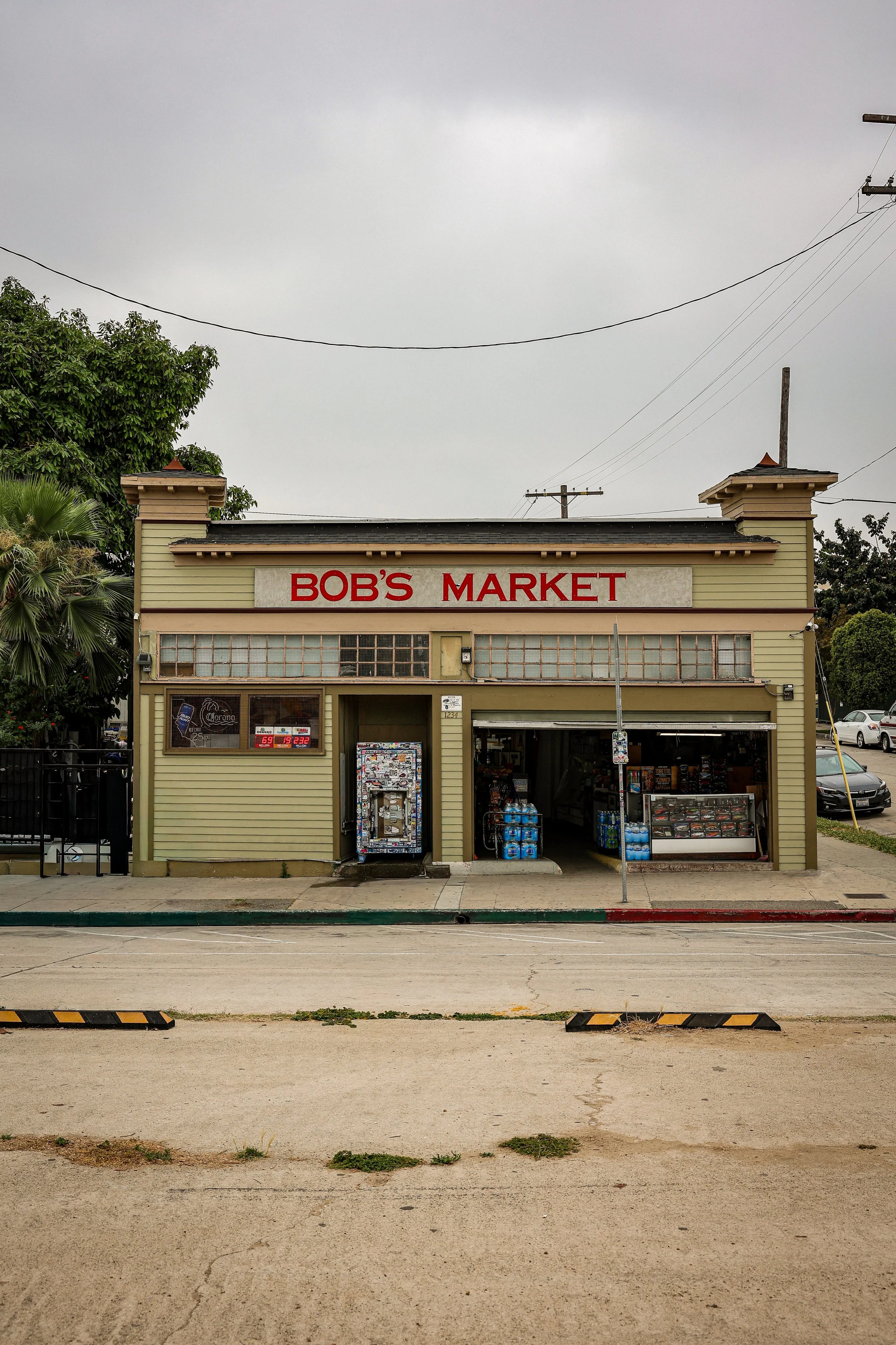 Front view of Bob's Market, a small grocery store with a beige exterior, large window, and open entrance displaying snacks and bottled water. There are parking spaces in front, power lines above, and a cloudy sky.
