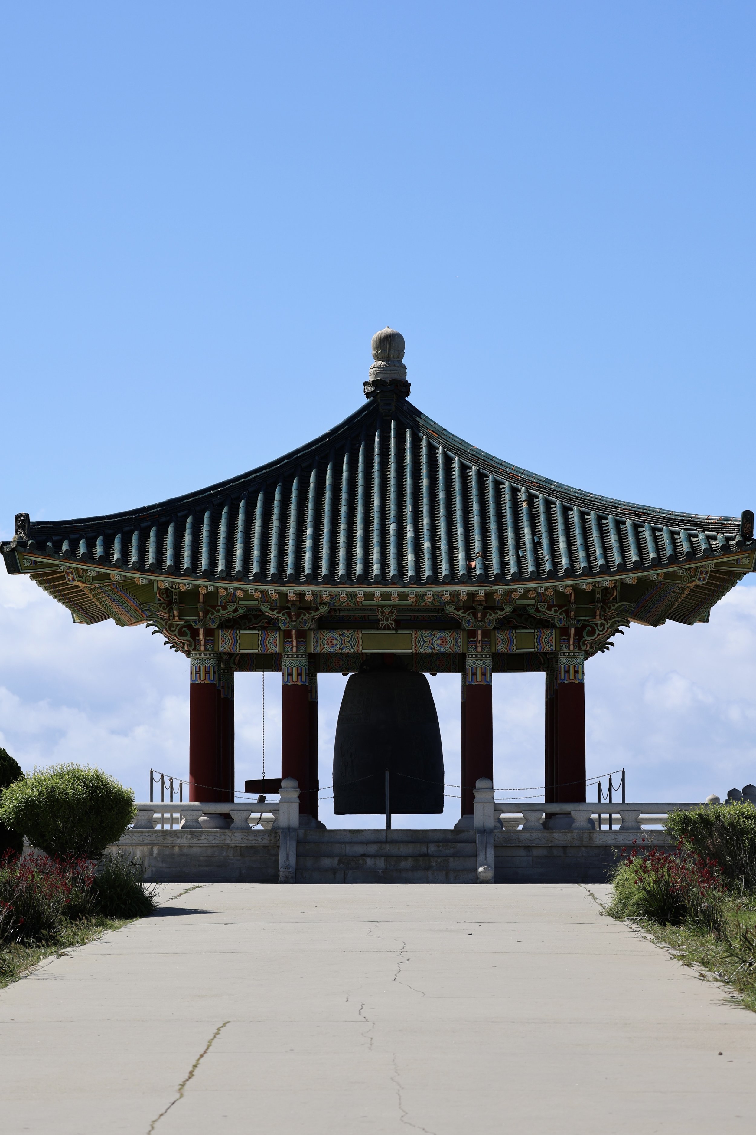 A traditional Korean bell pavilion with a large bronze bell beneath a curved, ornate roof with intricate patterns, set against a blue sky with some clouds and surrounded by greenery.