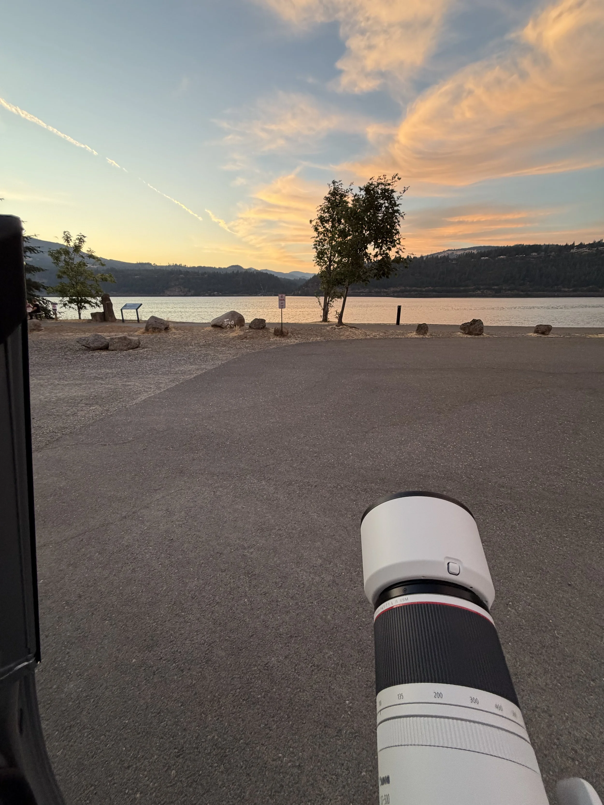 A sunset over a body of water with mountains in the distance and trees on the shore, viewed from a parking area with rocks, a sign, and a camera lens in the foreground.