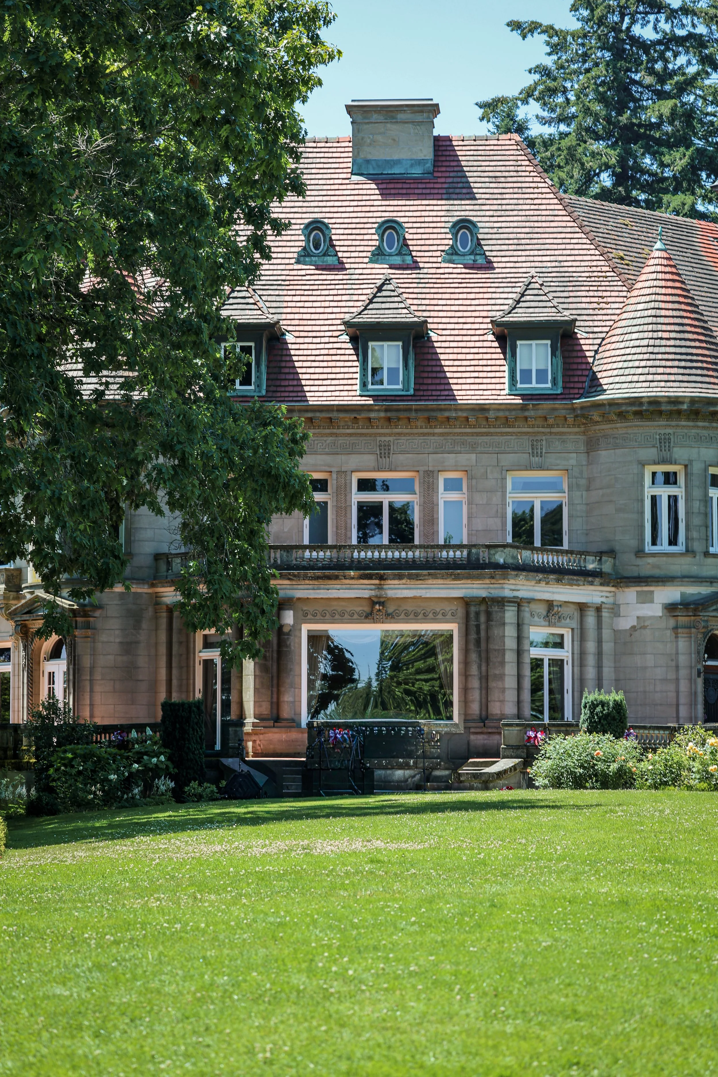 A large, elegant house with a stone facade and a red tiled roof. The house has multiple small dormer windows and a turret on the right. There is a well-maintained garden with colorful flowers in front, a lush green lawn, and tall trees surrounding the house.