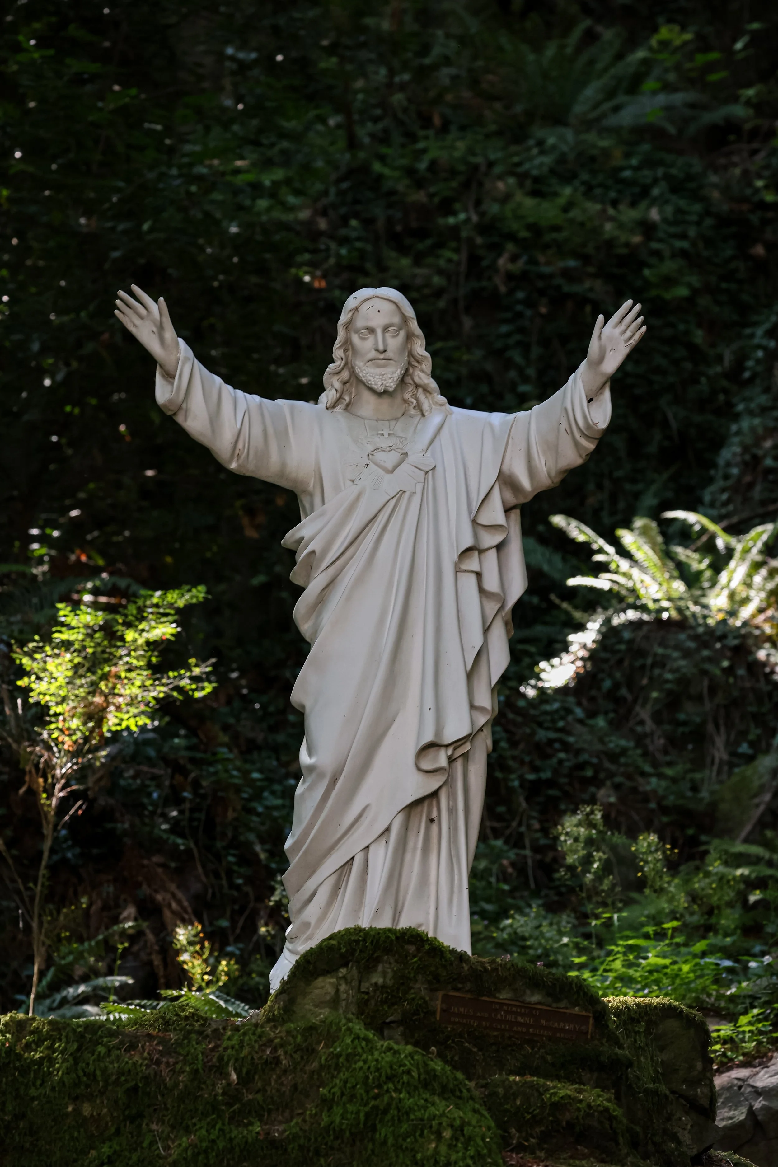 White statue of Jesus Christ with open arms, surrounded by lush greenery and moss-covered rocks in a forest setting.