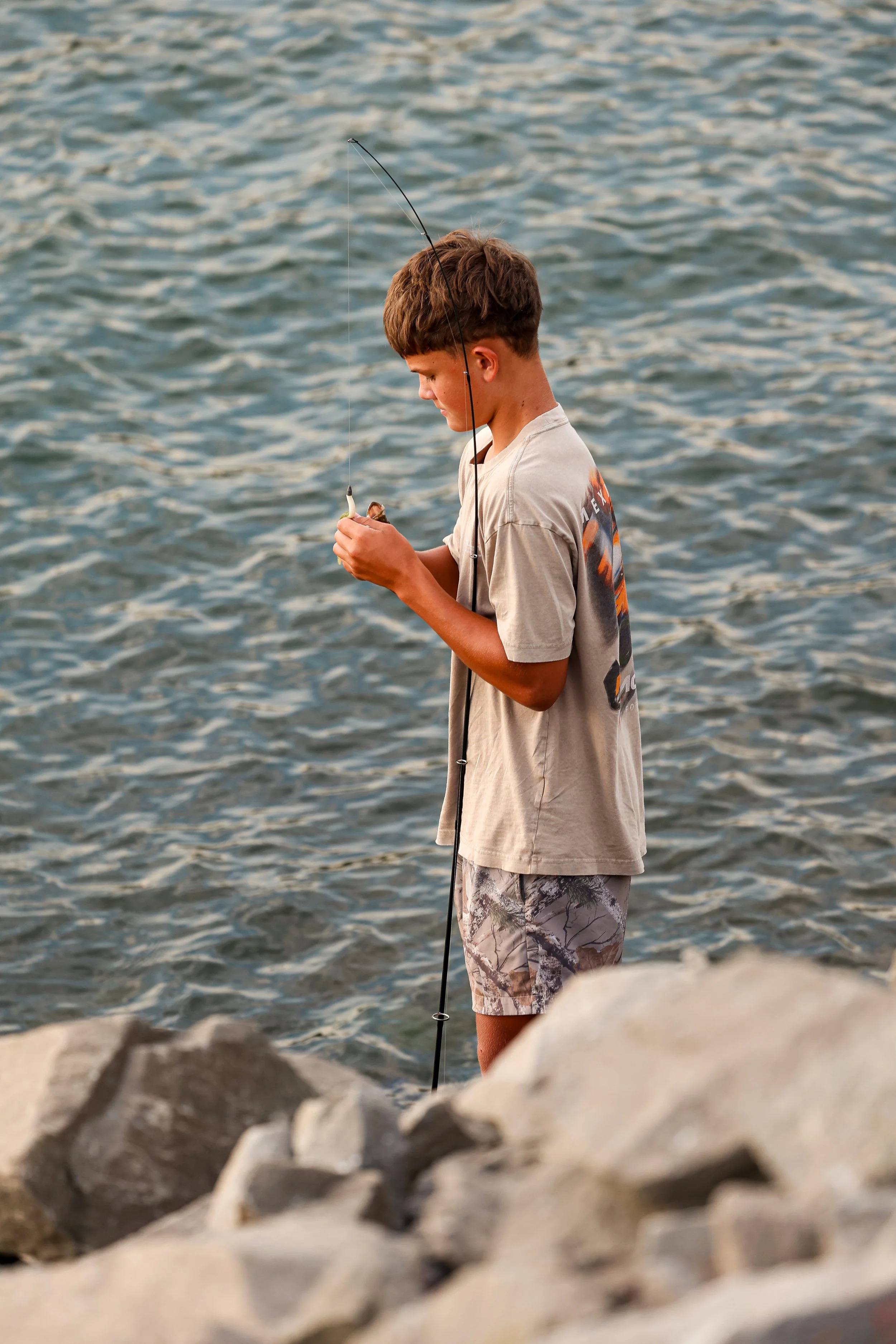 A teenage boy fishing by the water, wearing a beige T-shirt and camouflage shorts, with a fishing rod in his hands, standing near a rocky shoreline.