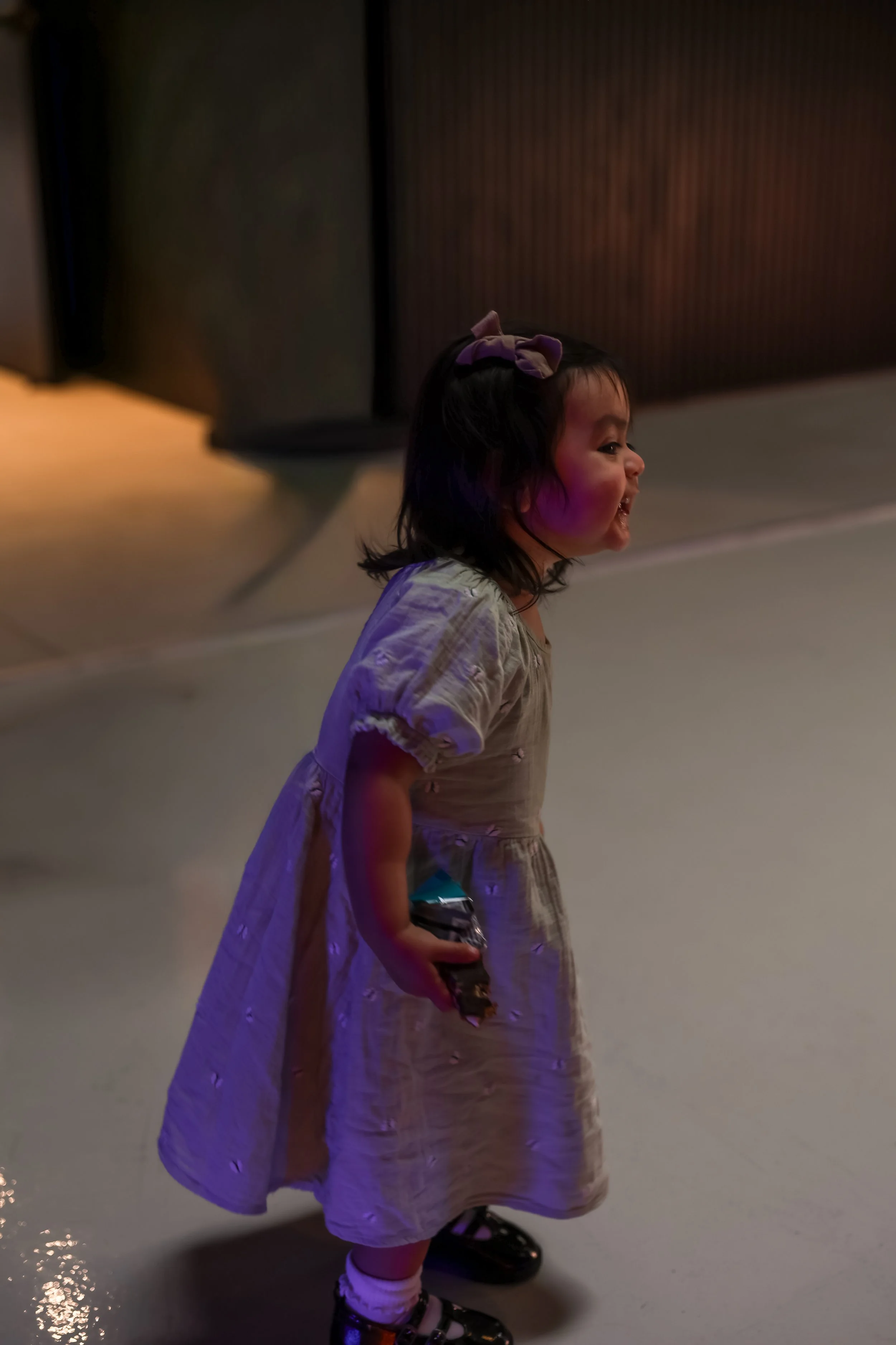 A young girl with dark hair and a purple bow in her hair, smiling and holding a small item, standing on a brightly lit floor with a dark, patterned wall in the background.