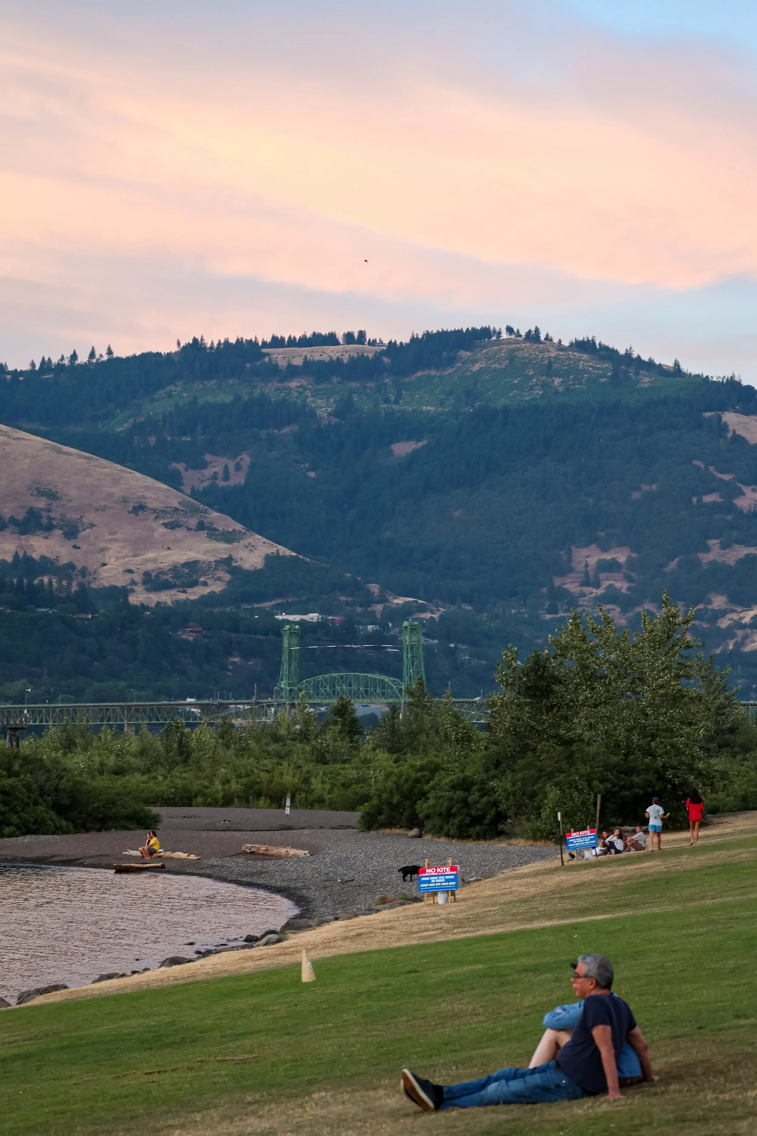 People sitting and relaxing along a river or lake with hills and mountains in the background, during sunset or dusk.