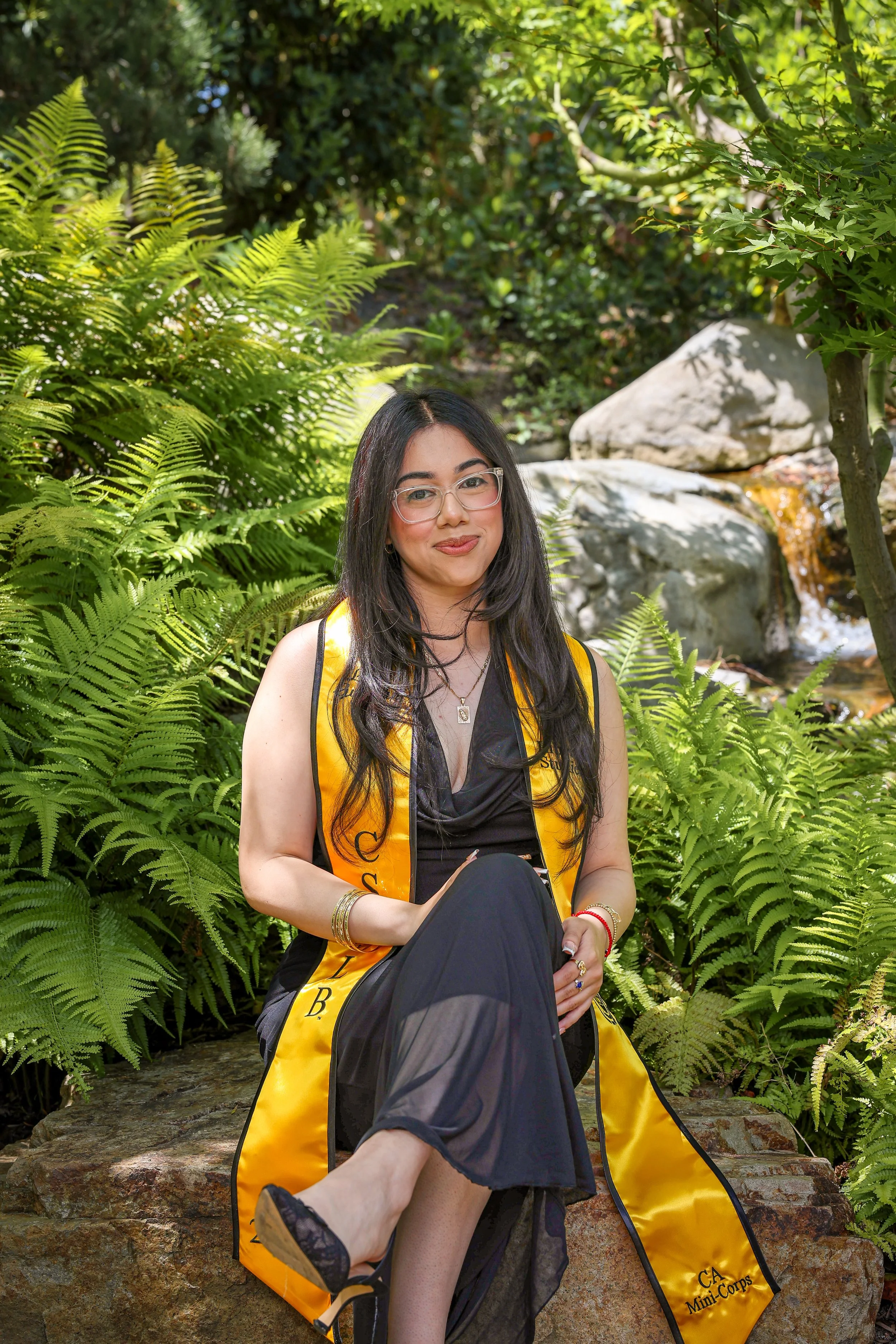 Young woman in a black dress and glasses sitting on a rock, wearing a yellow sash that reads CA Mini-Con, surrounded by lush green ferns, trees, and rocks in a garden or park.