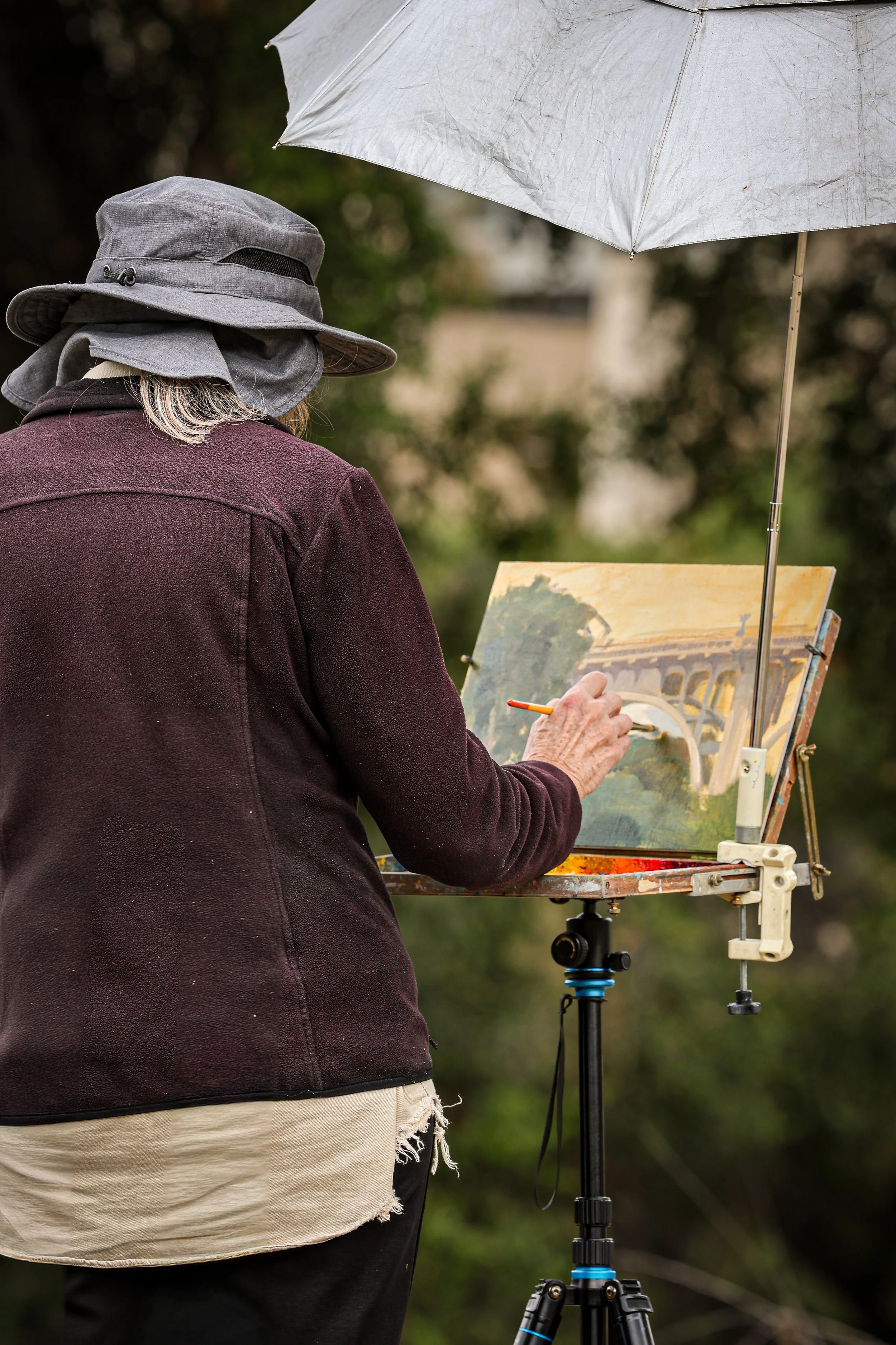 An artist painting outdoors on an easel with a cloudy white umbrella for weather protection, wearing a gray wide-brimmed hat, a dark red coat, and beige and black clothing.