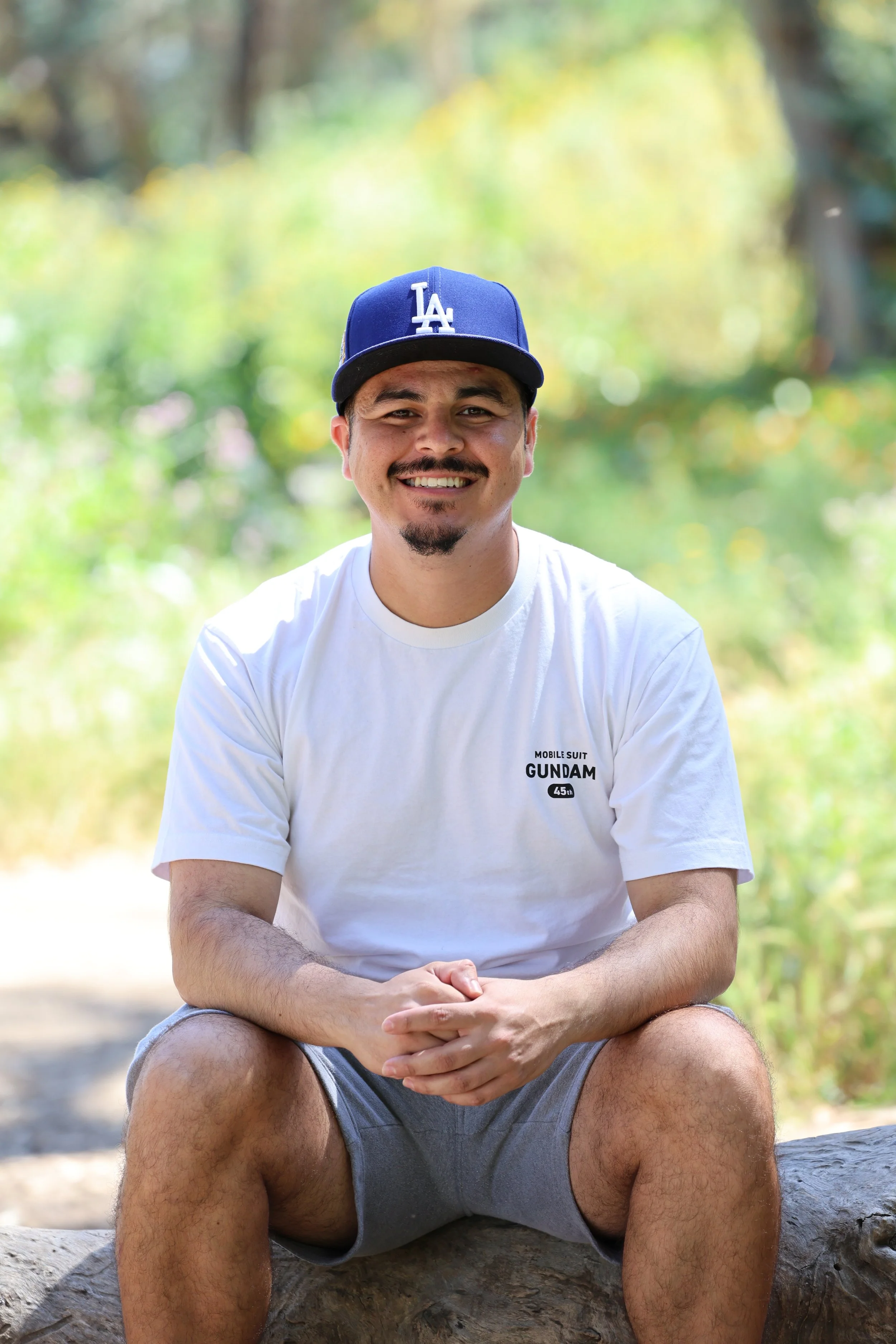A man sitting on a large fallen tree trunk outdoors, smiling, wearing a blue Los Angeles Dodgers baseball cap and a white t-shirt with a small logo, with greenery in the background.