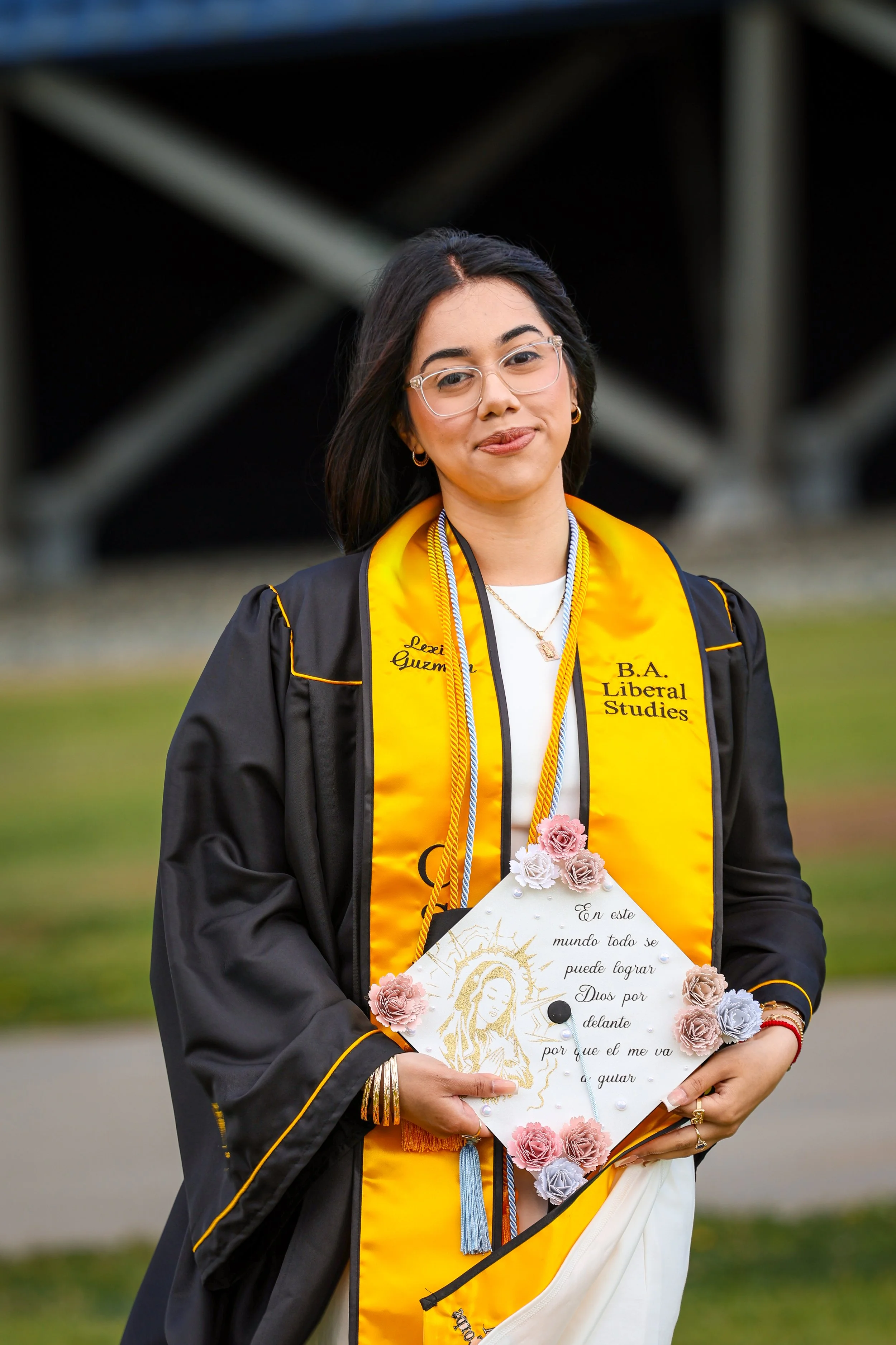 A young woman dressed in a graduation gown and stole, holding a decorated certificate or diploma, standing outdoors with a blurred background of structural beams and greenery.