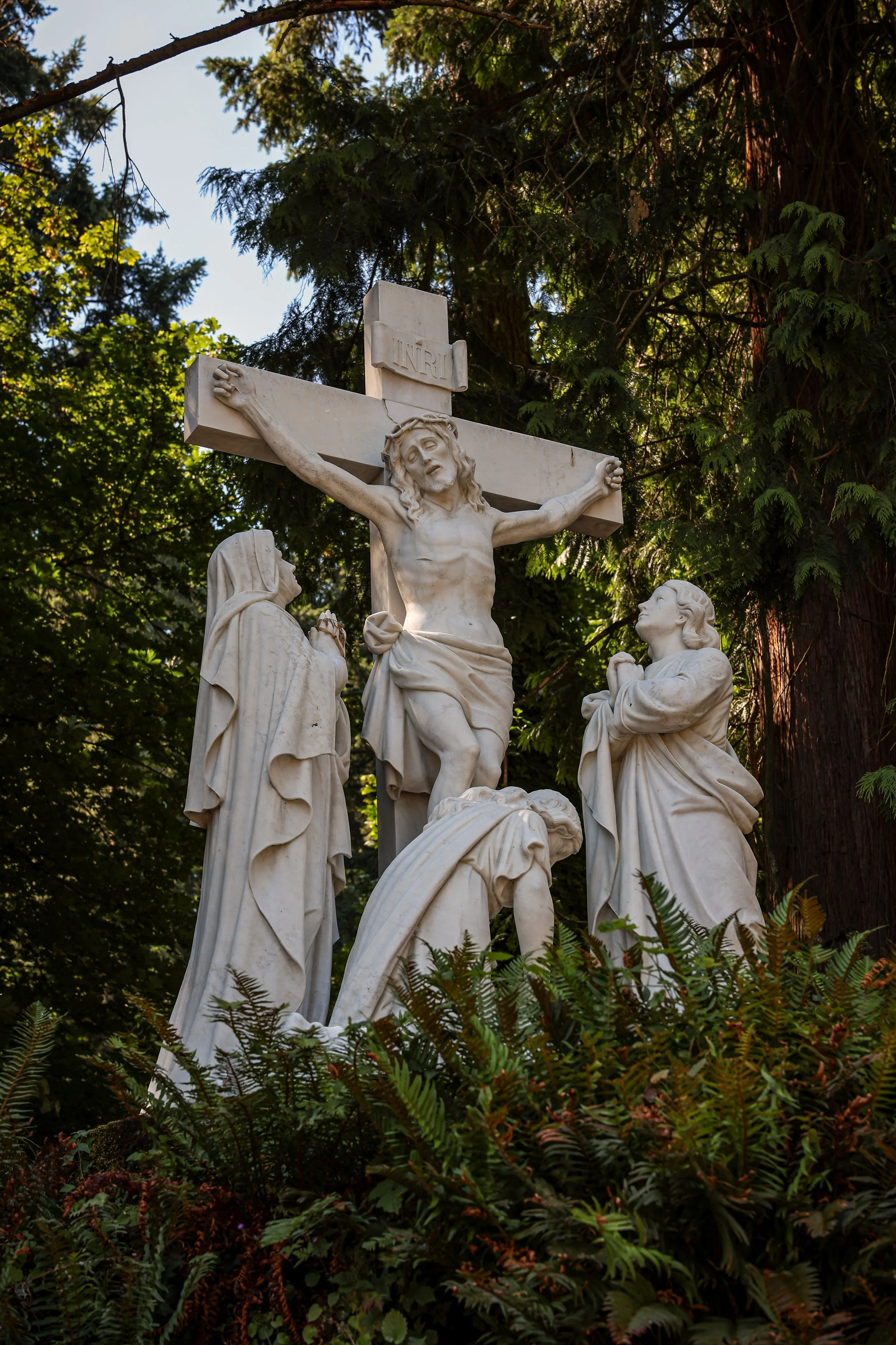 A white marble religious sculpture depicting Jesus Christ on the cross, with Mary and another woman at his feet, surrounded by green trees and plants.