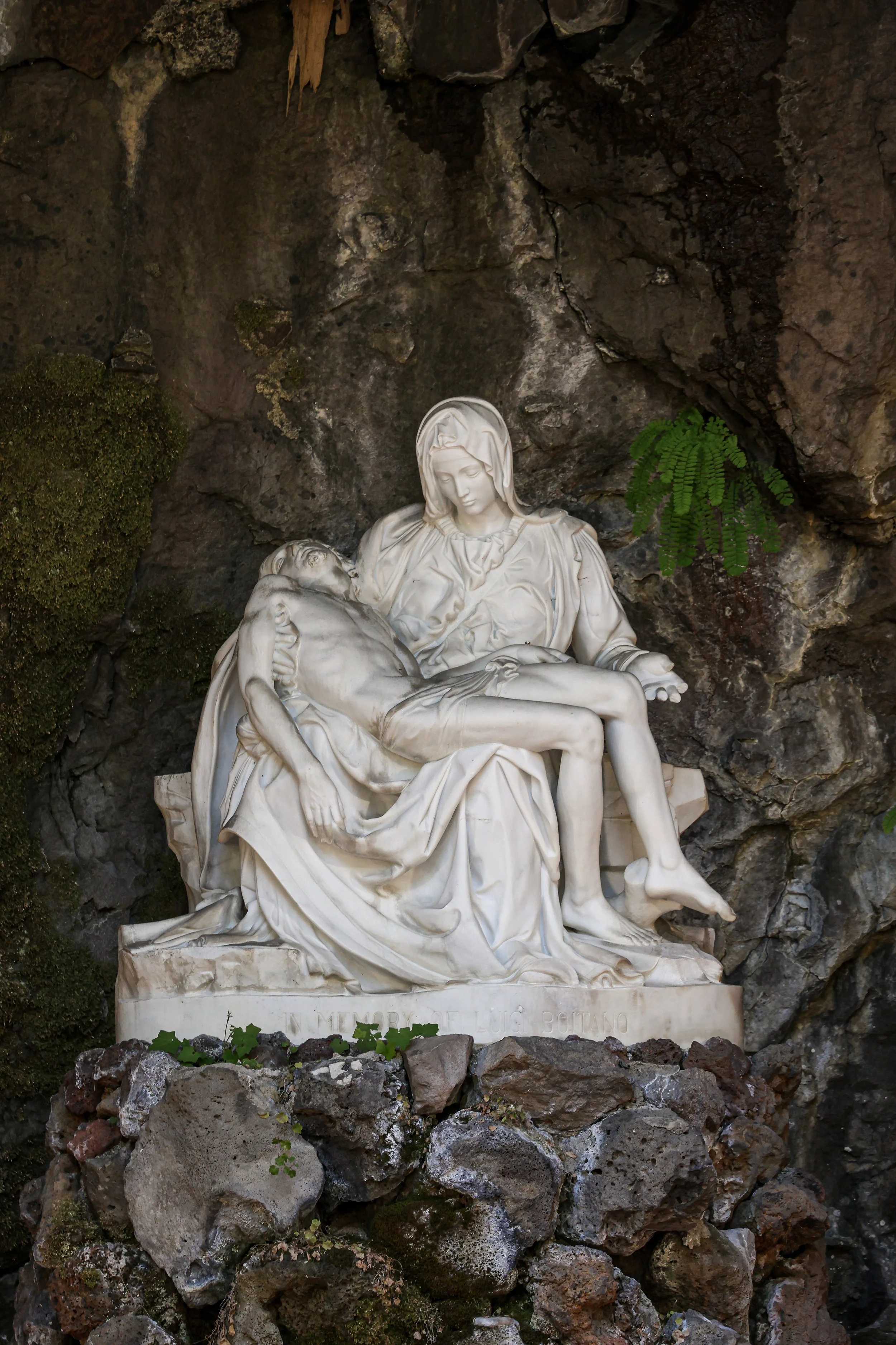 A white marble sculpture of the Pieta, depicting Virgin Mary holding the body of Jesus Christ after his crucifixion, placed on a rocky background with some greenery.
