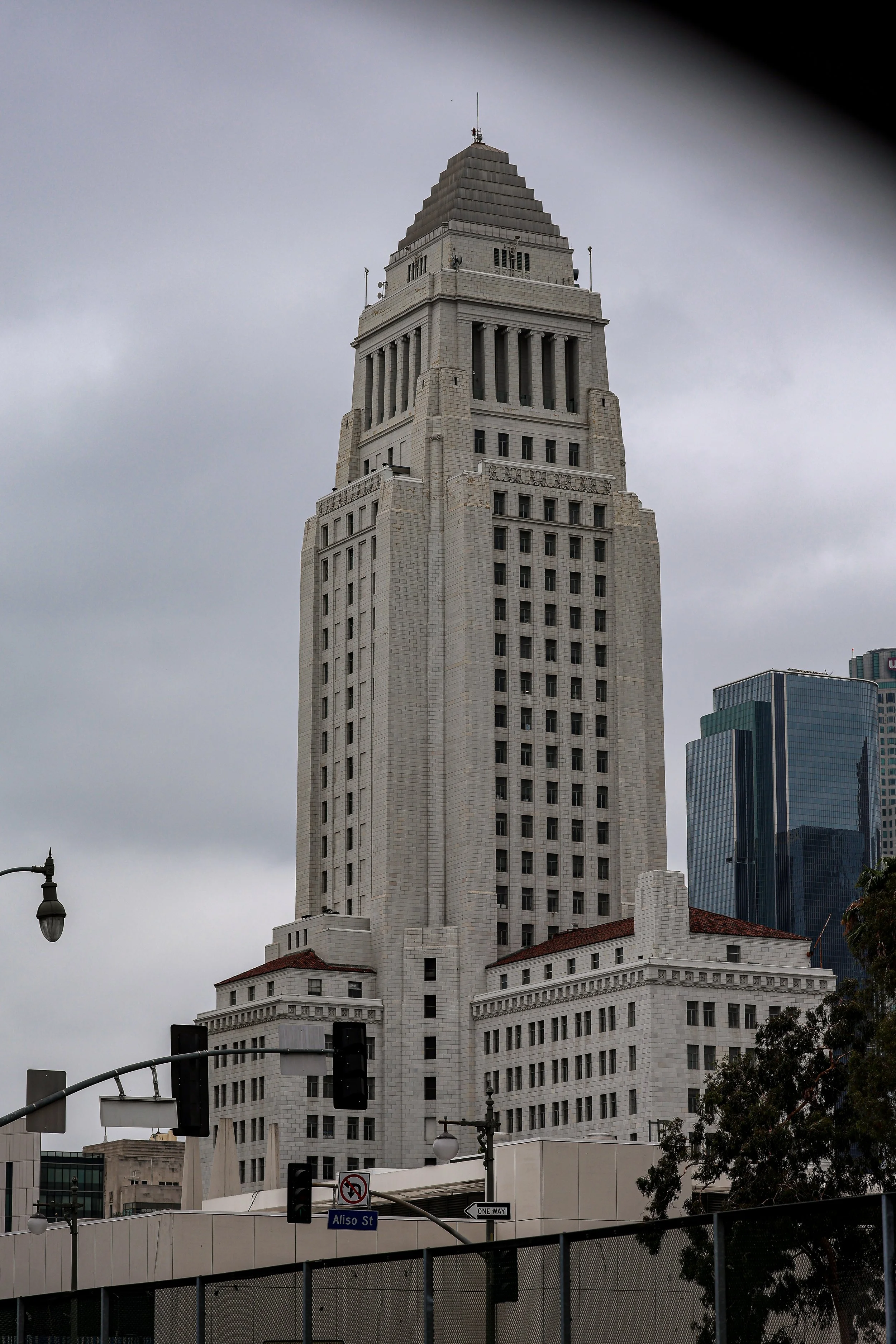 Tall, white building with an Art Deco architectural style, featuring a stepped, pyramid-shaped top, against a cloudy sky. In the foreground, traffic lights, street signs, and some trees are visible.