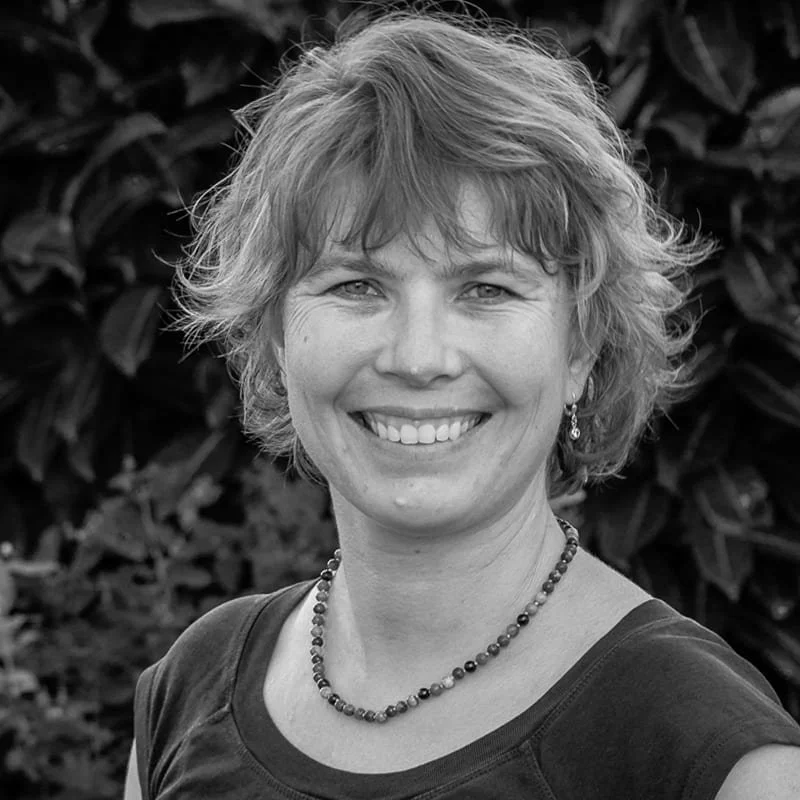 Schwarz-weiß-Fotografie einer lachenden Frau mit kurzen, gewellten Haaren, tragen Kette und Ohrringe, vor einem Hintergrund aus Blättern.
