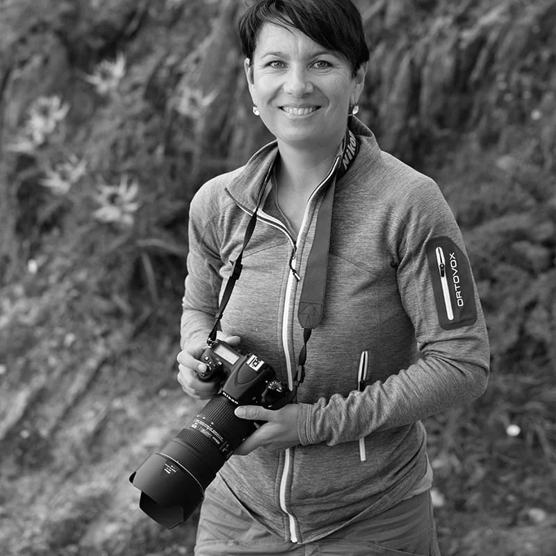Frau mit Kamera vor natürlicher Hintergrundscenerie, lächelnd, in sportlicher Kleidung, draußen.
