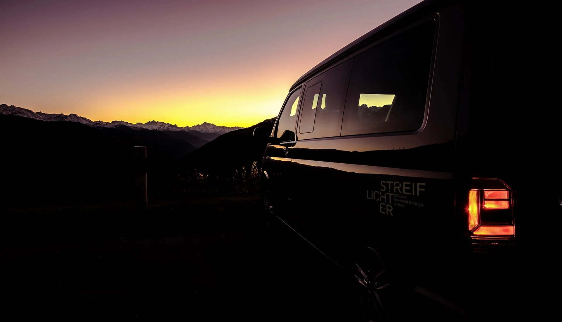 Ein schwarzer Van mit reflektierter Bergwelt im Fenster steht bei Sonnenuntergang oder -aufgang vor einer Bergkette.