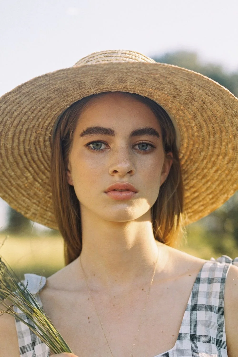 Young woman with blue eyes wearing a wide-brimmed straw hat and a checkered sleeveless dress outdoors.