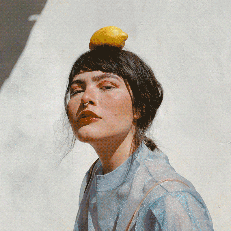 A young woman with dark hair and a septum piercing is outdoors under sunlight, balancing a lemon on her head, wearing a sheer, shimmery top.