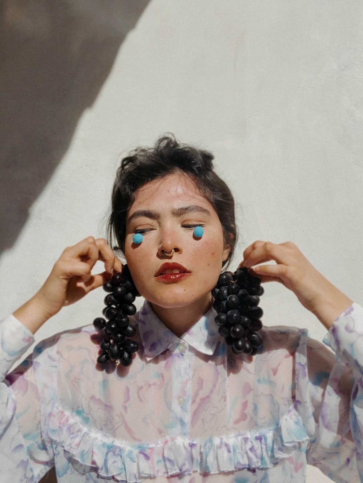 A woman with short dark hair, closed eyes, and red lipstick, is wearing a colorful, patterned shirt with ruffles. She has blue patches under her eyes, a septum piercing, and is holding large bunches of dark grapes on both sides of her head like earrings.