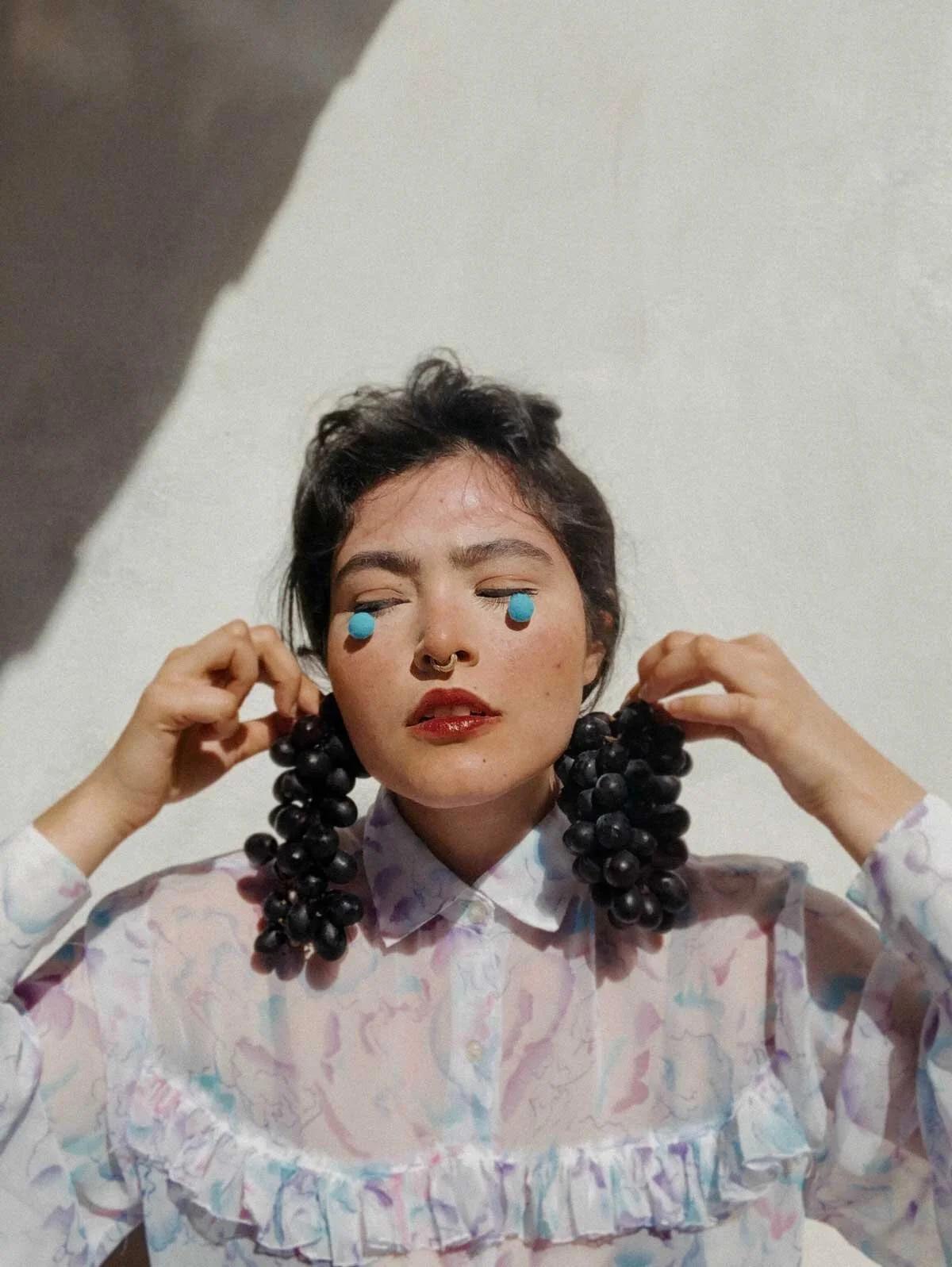 A woman with short dark hair and makeup, wearing blue cream earrings, holding oversized black grape earrings, and a white blouse with pastel water mark pattern, standing against a plain light-colored wall.