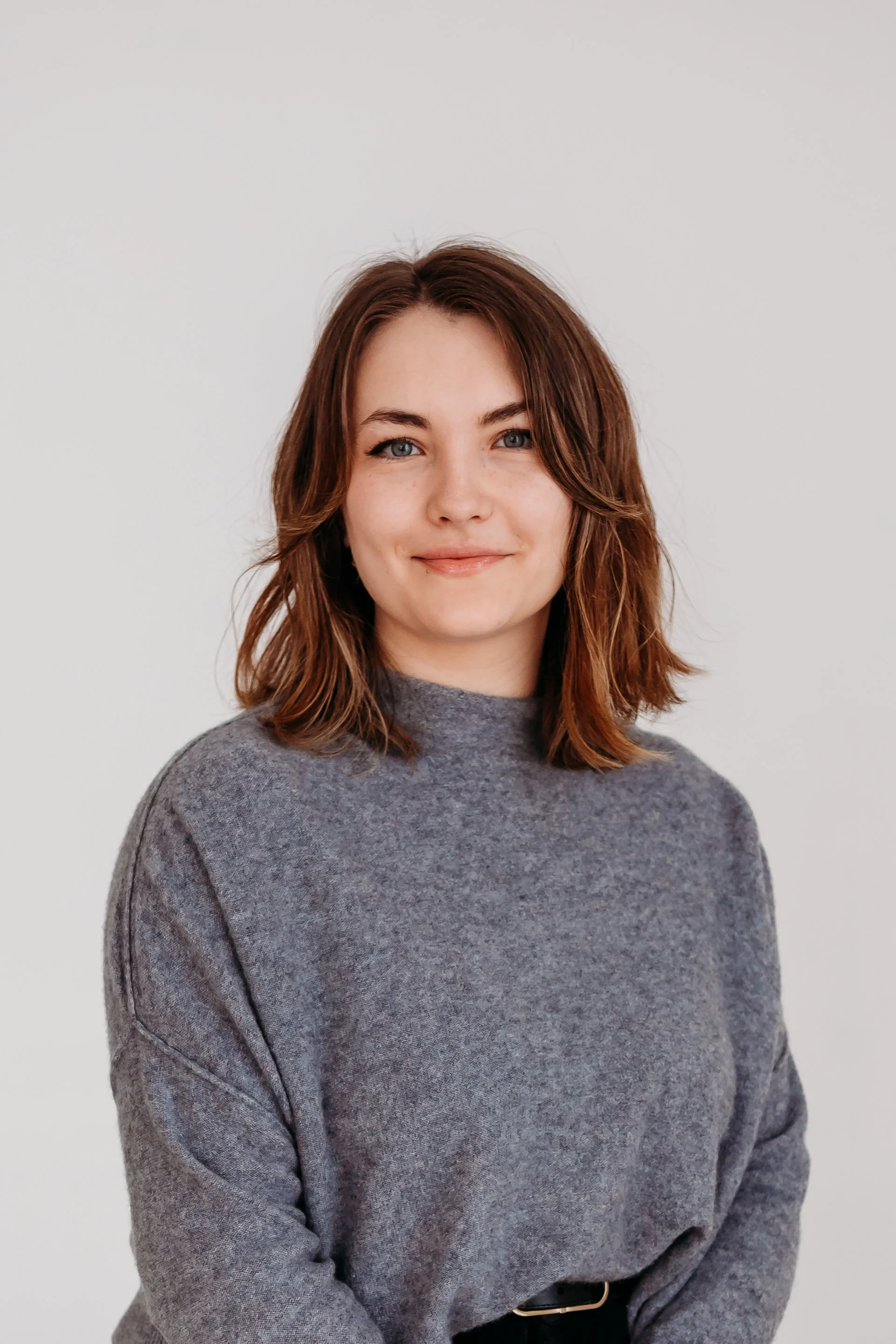 A woman with shoulder-length brown hair, blue eyes, wearing a gray sweater, smiling against a plain white background.