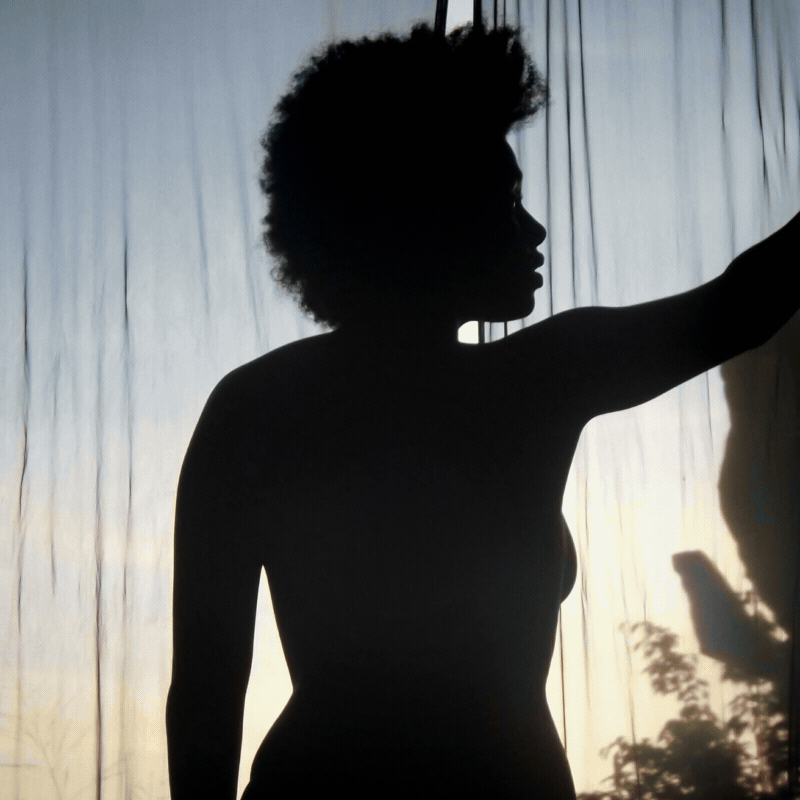 Silhouette of a woman with natural hair in front of a window with curtains, backlit by daylight.