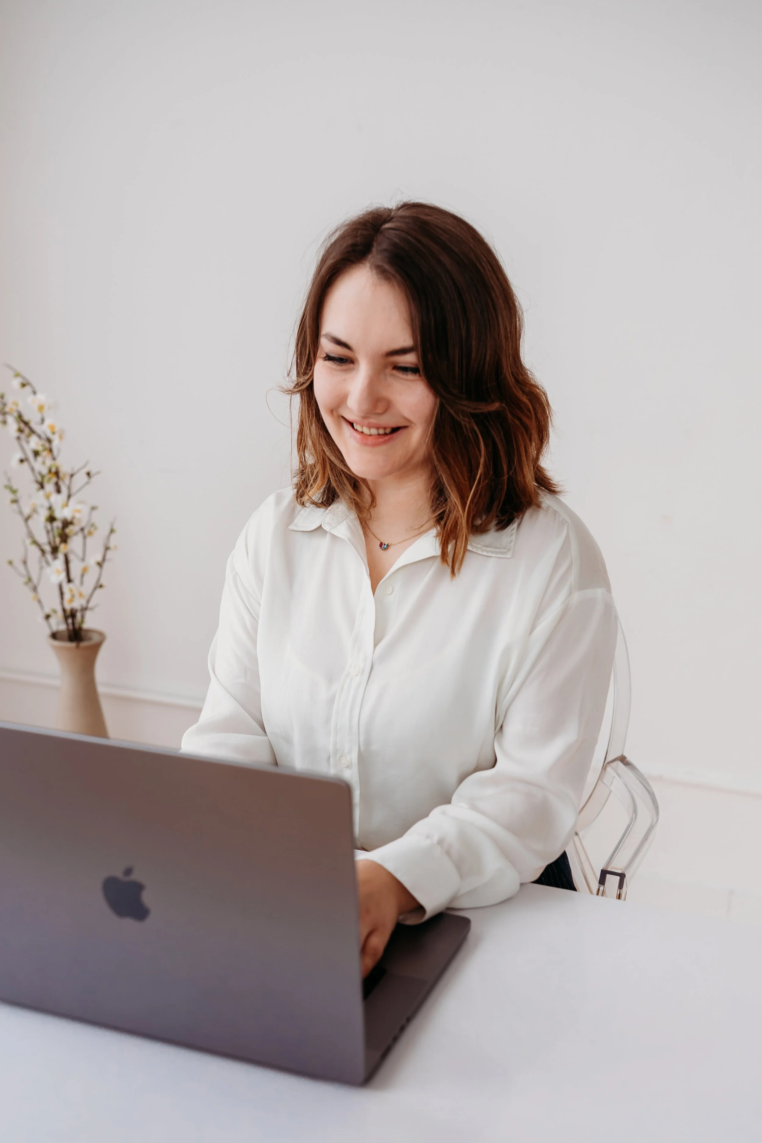 A woman with shoulder-length brown hair smiling while working on a laptop in a minimal white office space with a pot of white flowers on the side.