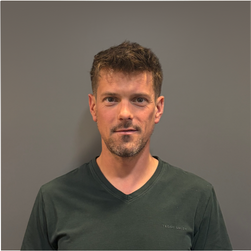 Portrait d'un homme jeune avec des cheveux courts et bruns, portant un t-shirt vert foncé, debout devant un fond gris.