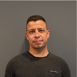 Portrait d'un homme avec une expression neutre, portant un pull noir, devant un mur gris