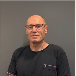 Homme chauve portant des lunettes et un t-shirt noir, posé contre un fond gris.
