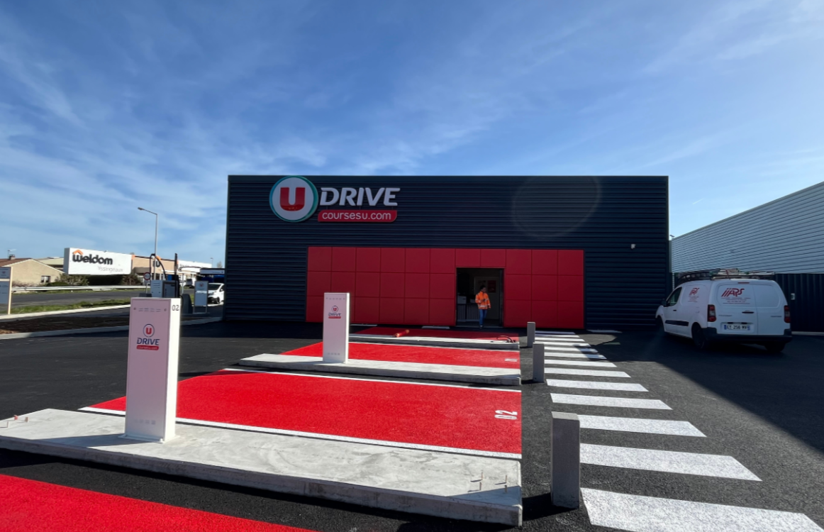 Entrée d'une station de drive avec des places de stationnement rouges, panneau avec le logo 'U DRIVE' et un véhicule blanc garé à droite.