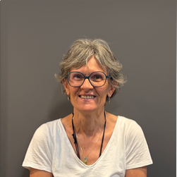 Femme âgée avec des cheveux gris, portant des lunettes et un t-shirt blanc, souriant debout devant un mur gris.